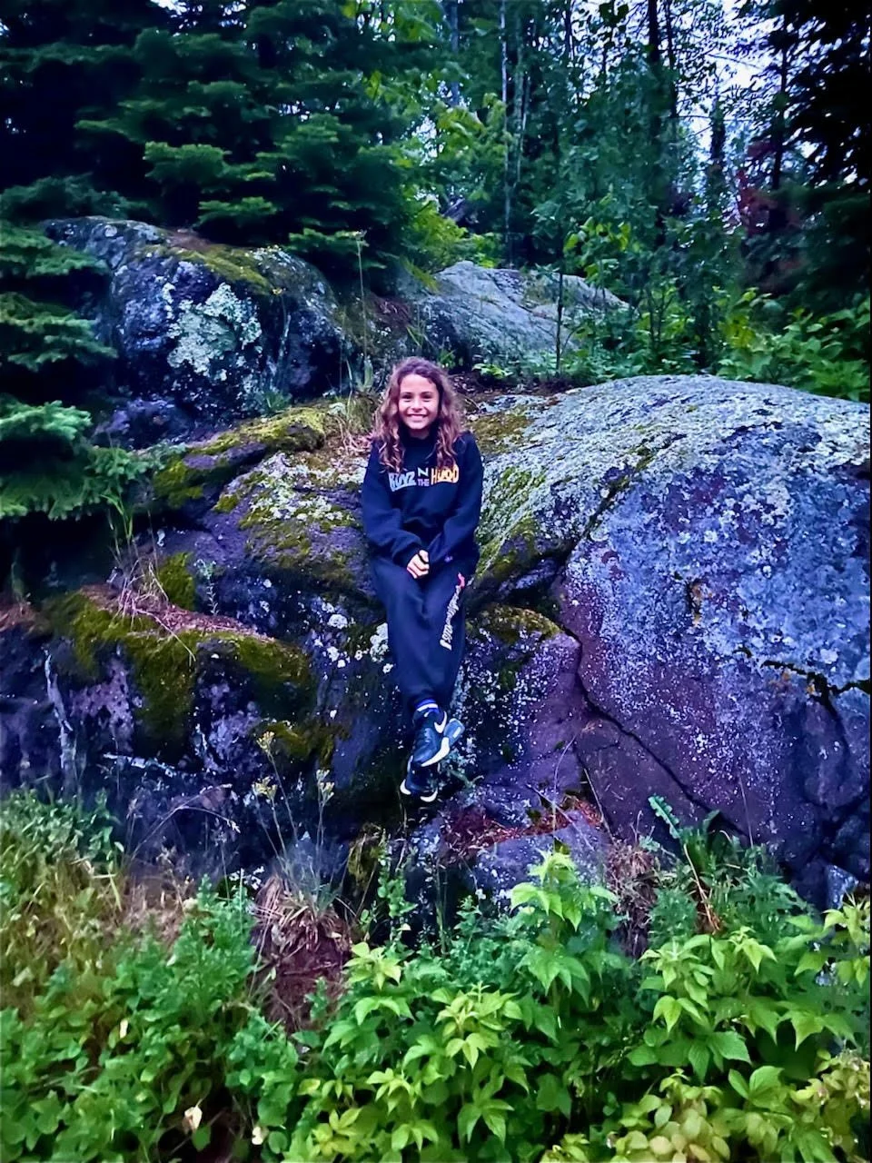 A young girl smiling and sitting on a moss-covered rock in a lush green forest surrounded by trees and plants.