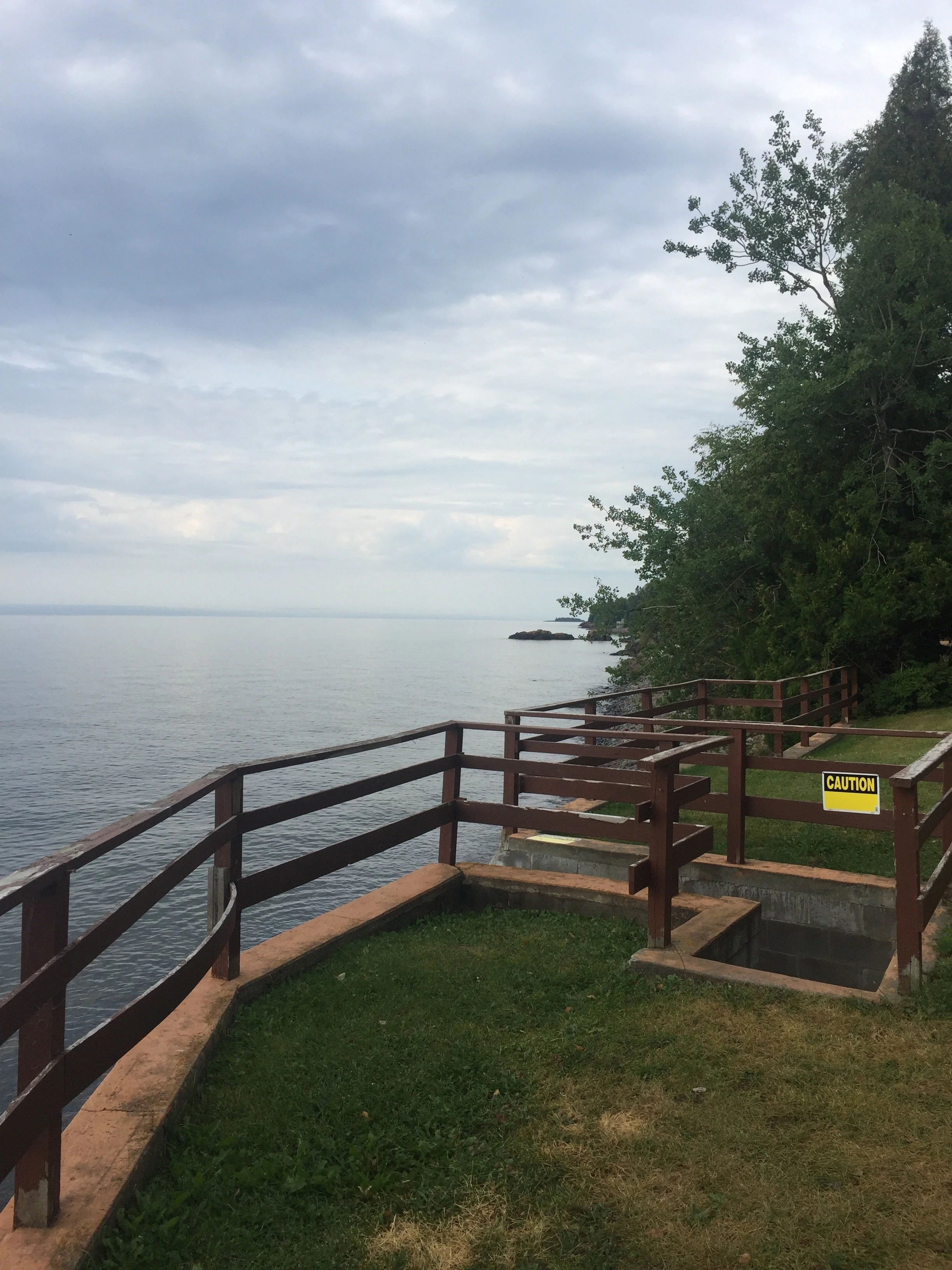 A lakeside walkway with a wooden railing, green grass, and trees on the right side. The sky is cloudy and there is a caution sign on the railing.