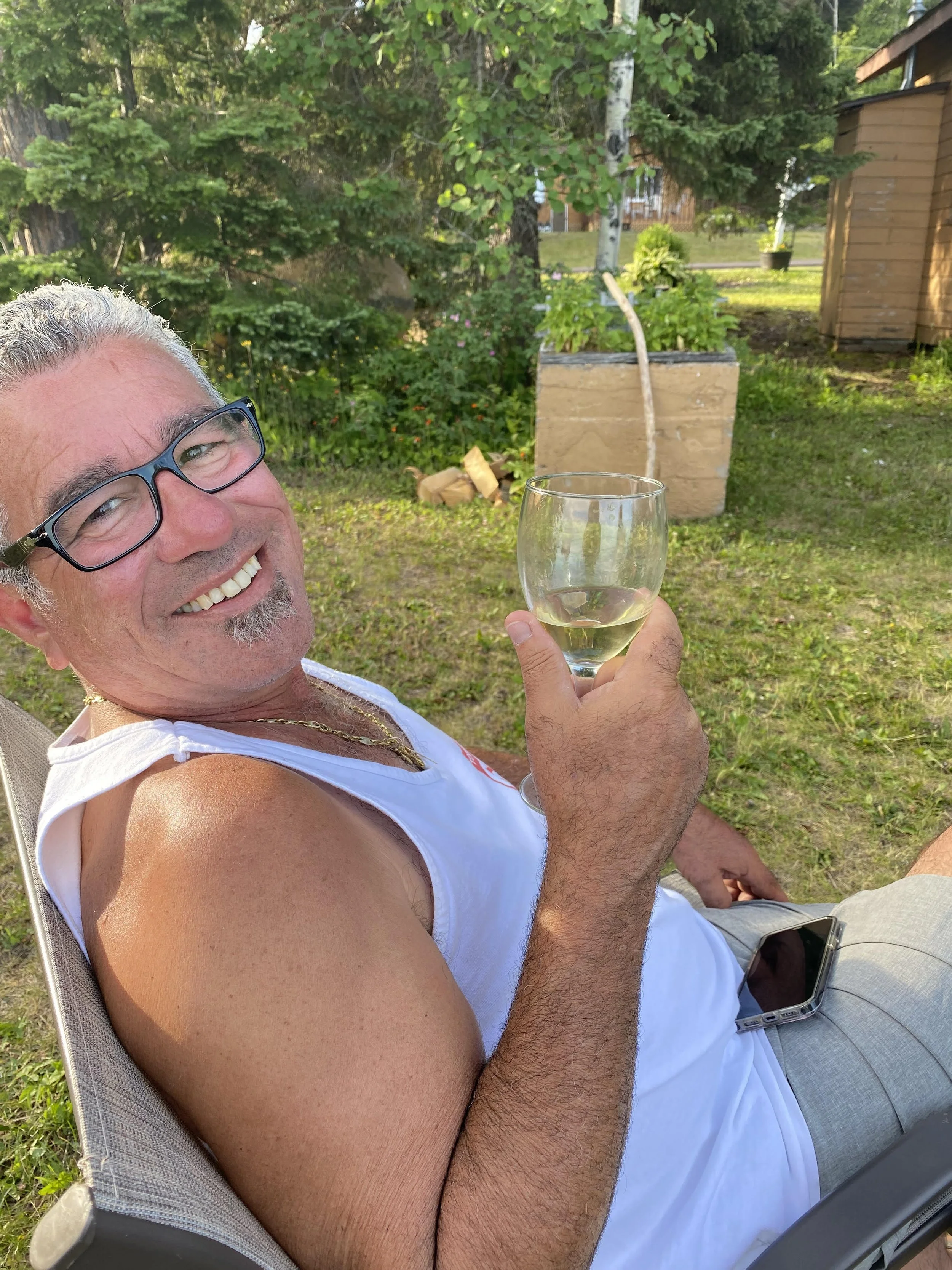 A middle-aged man with gray hair, glasses, and a goatee, smiling and holding a glass of white wine, relaxing outdoors in a garden.