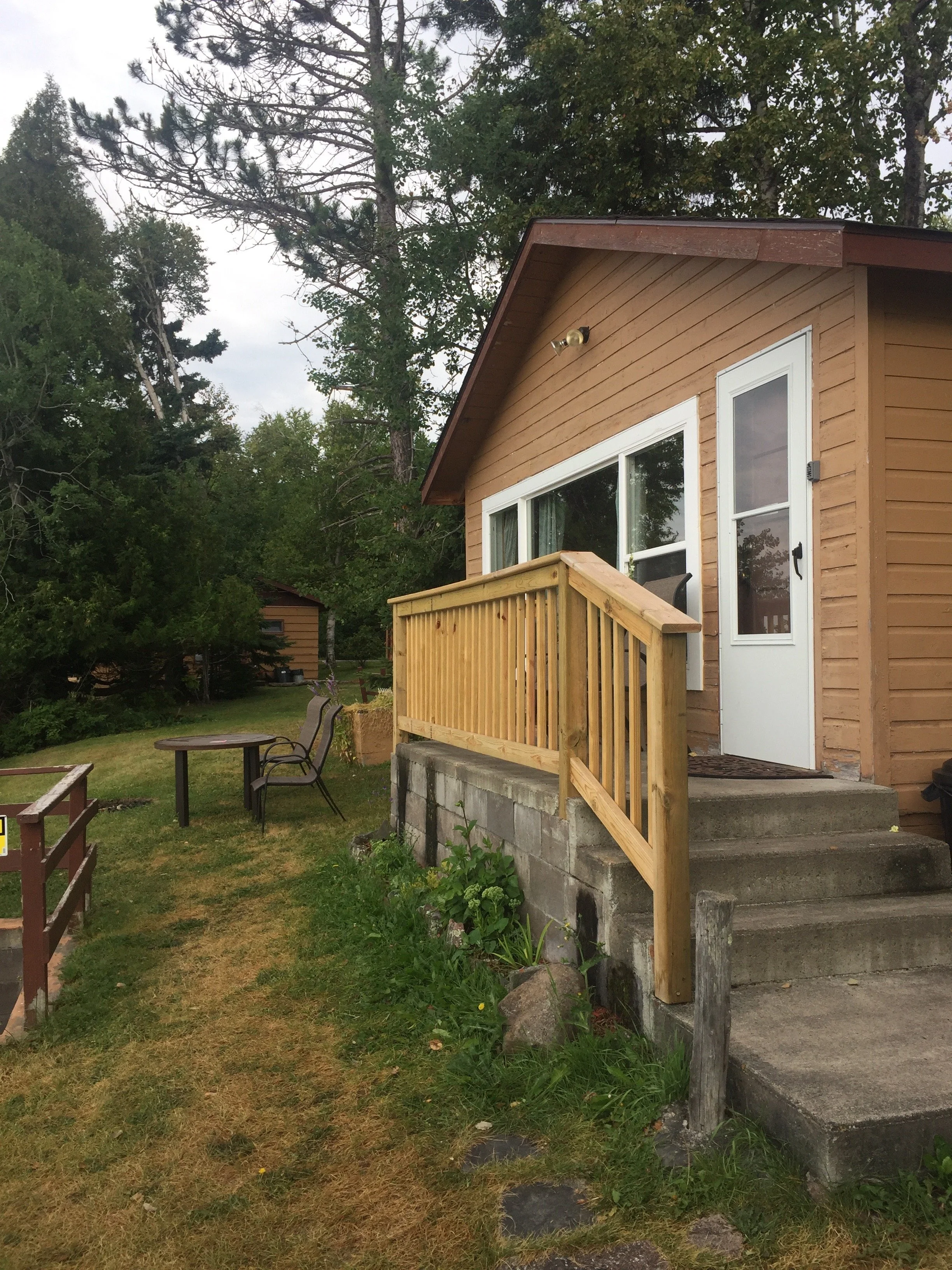 A small wooden house with a front door and a window, a steps leading up to a porch with a wooden railing, and a garden with chairs and a table in the yard, surrounded by green trees under cloudy sky.