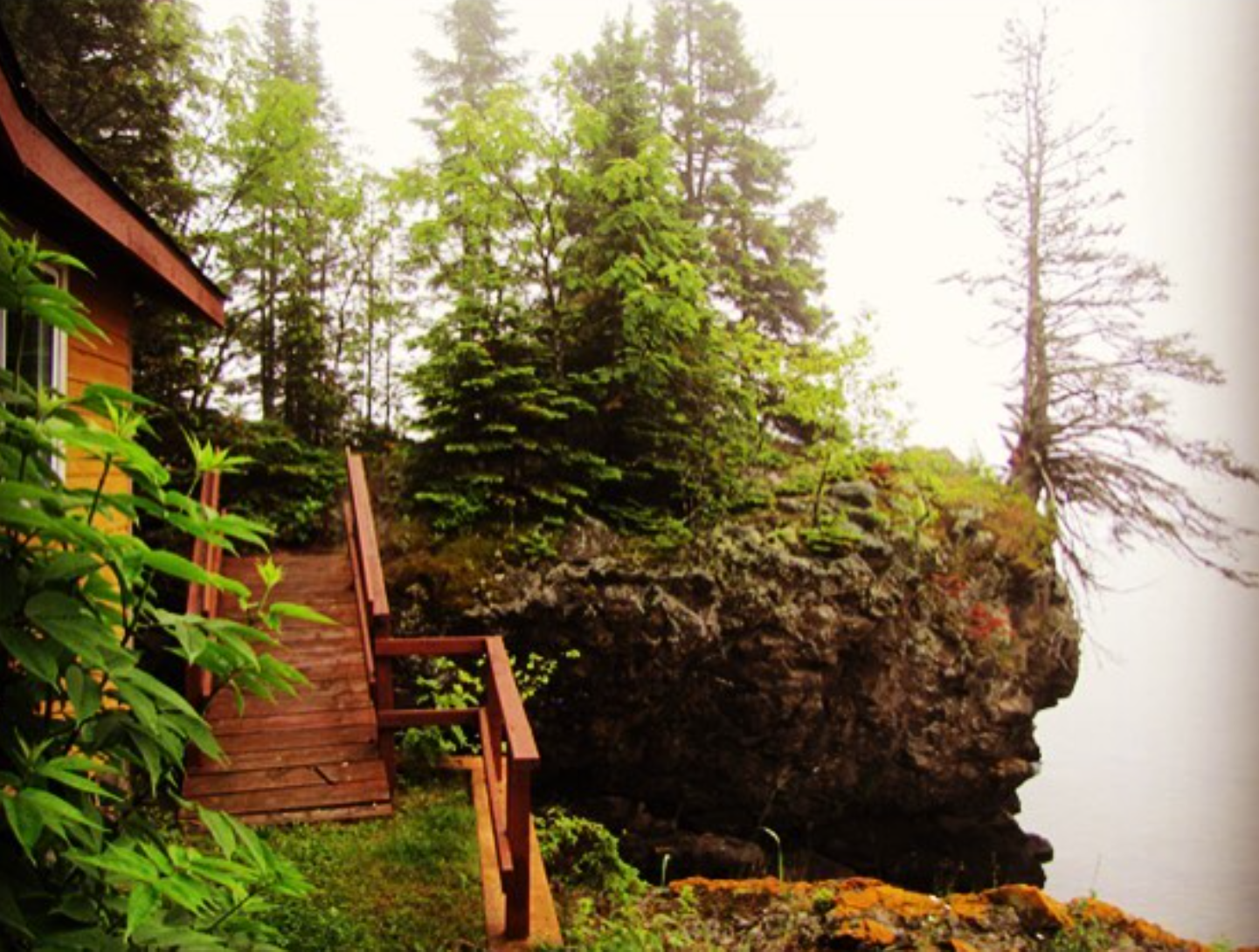A wooden staircase leading down from a house on the left side of the image to a rocky, forested cliffside with trees and mossy rocks, overlooking a body of water.