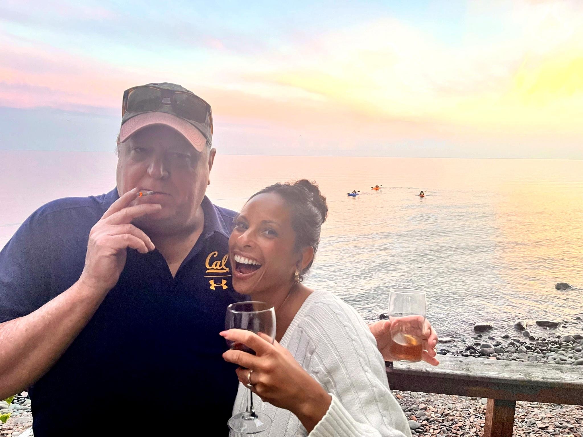A man and woman smiling on a beach at sunset, holding drinks, with kayakers on calm water in the background.