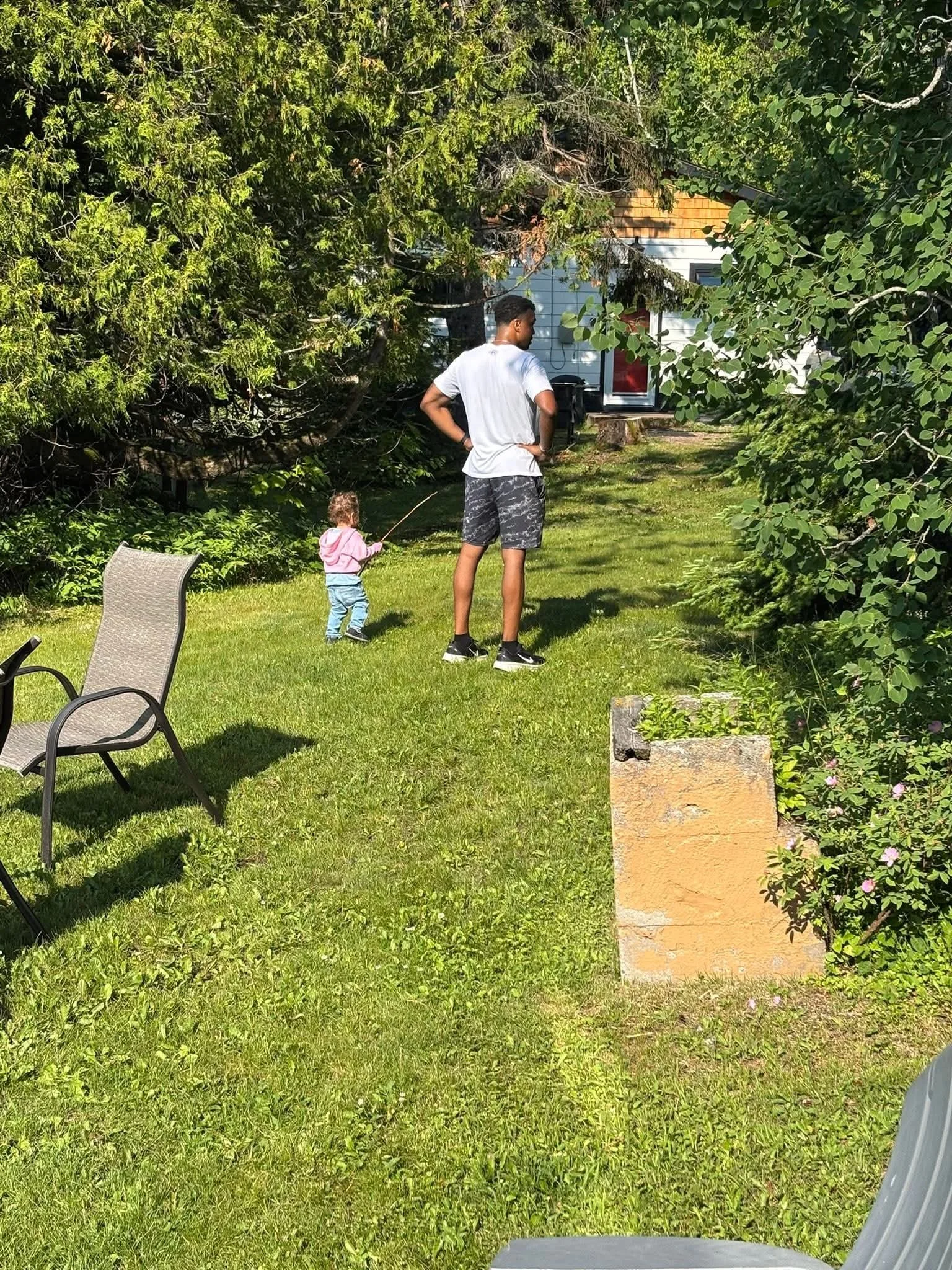 A man and a young girl, possibly his daughter, are in a backyard with green grass and trees. The girl is playing with a stick, and the man is standing with his hands on his hips, facing away from the camera. There are outdoor chairs, a house in the b