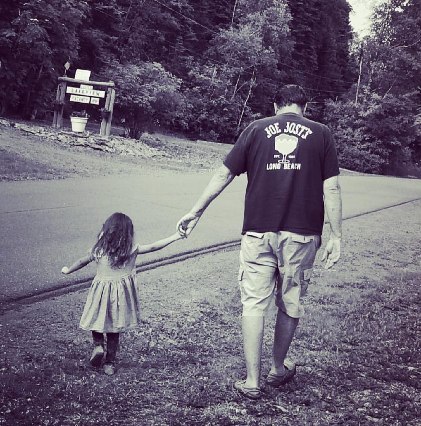 A black-and-white photo of a man and a young girl walking hand in hand on a grassy roadside. The man is wearing a t-shirt with the words 'Joe Jost's Long Beach' and appears to be guiding the girl, who is wearing a dress, away from a sign that reads '