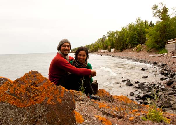 Two people sitting on rocks by a lakeside, smiling, with trees along the shore in the background.