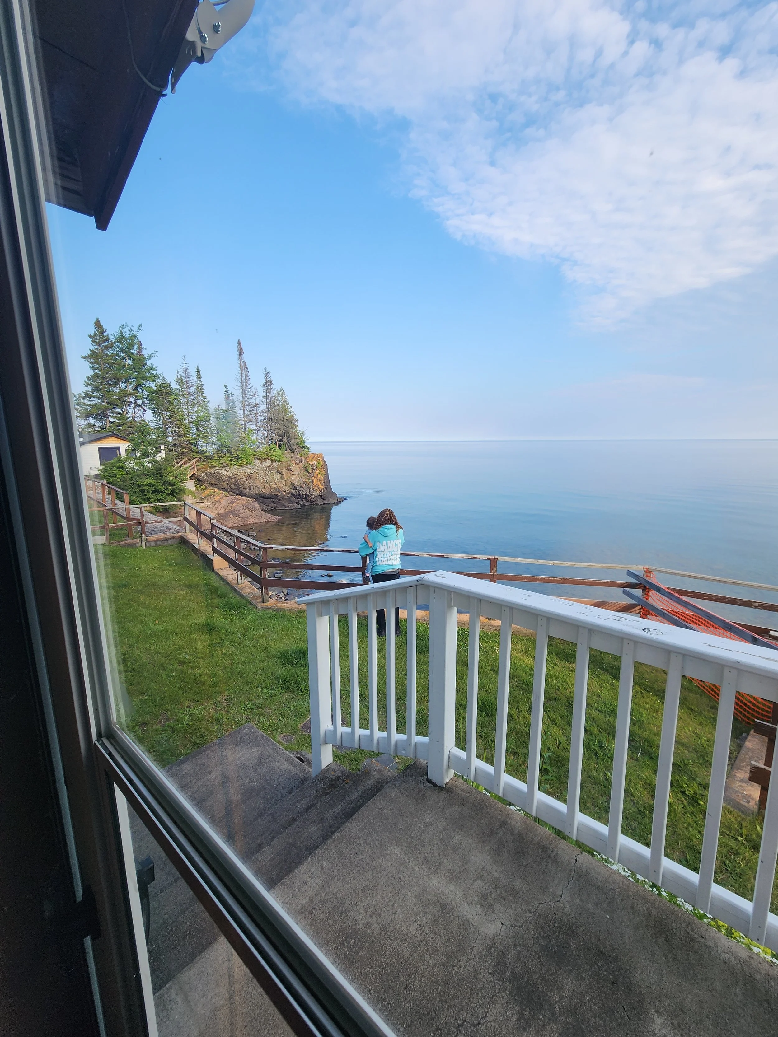 View of a woman standing on a grassy lawn near a wooden railing, looking out at a calm body of water with a small rocky landmass covered in trees, seen through a window with white fencing and stairs in the foreground.
