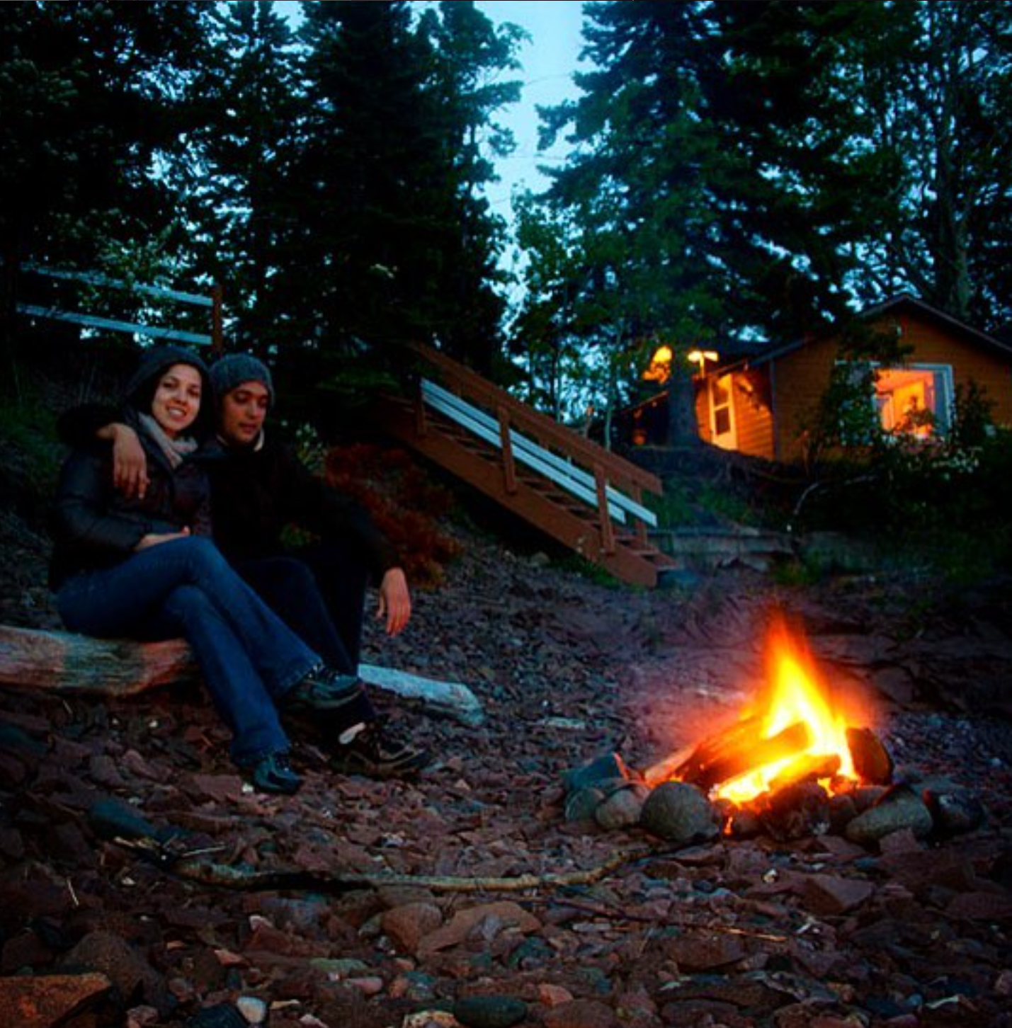 Two people sitting by a campfire on a rocky ground at dusk, with trees and a house with warm lights in the background.