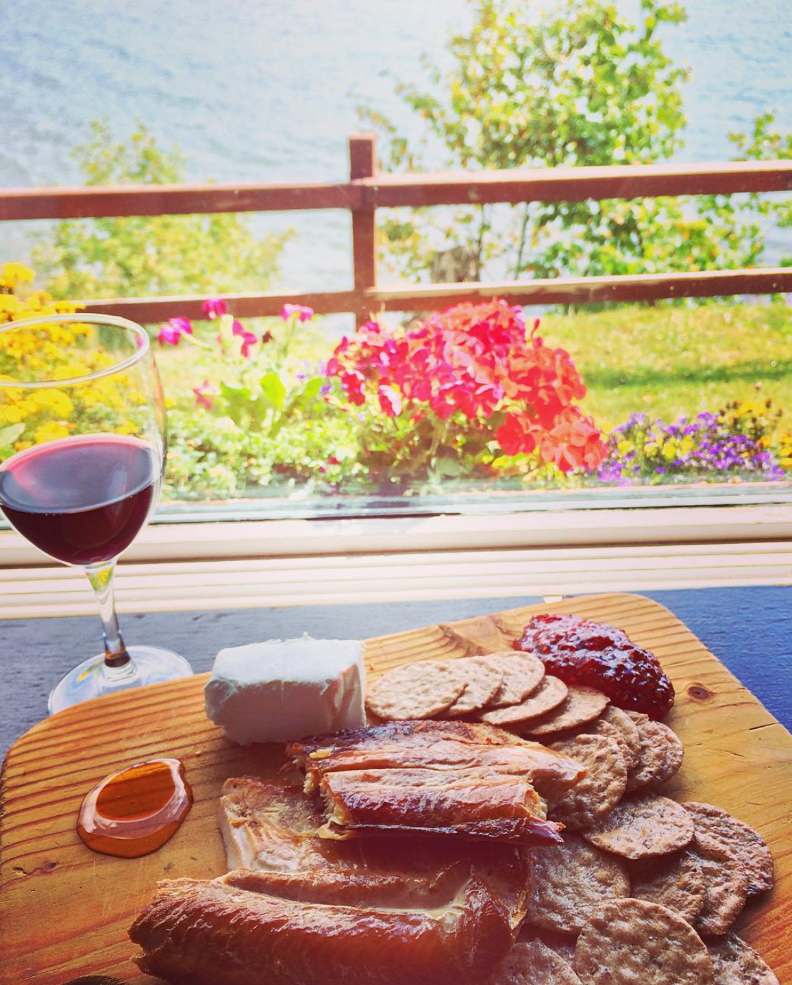 A cheese and meat board with a glass of red wine, placed on a windowsill overlooking a garden with colorful flowers and a body of water in the background.