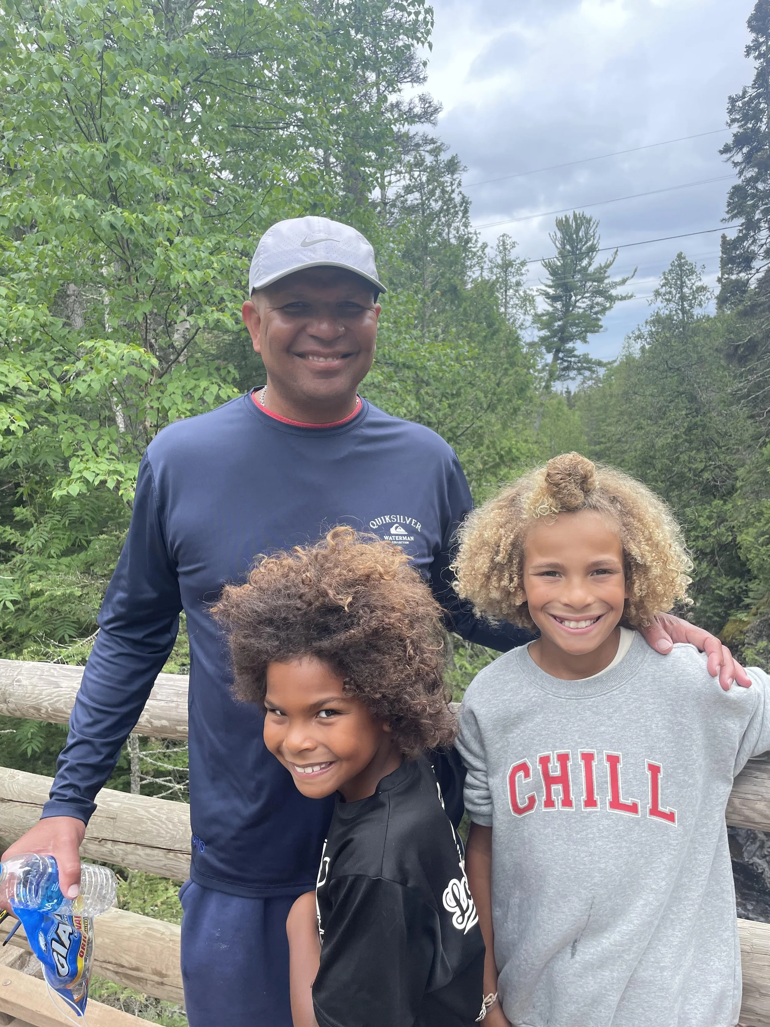 A man and two young girls outdoors on a wooden bridge, smiling with trees and cloudy sky in the background.