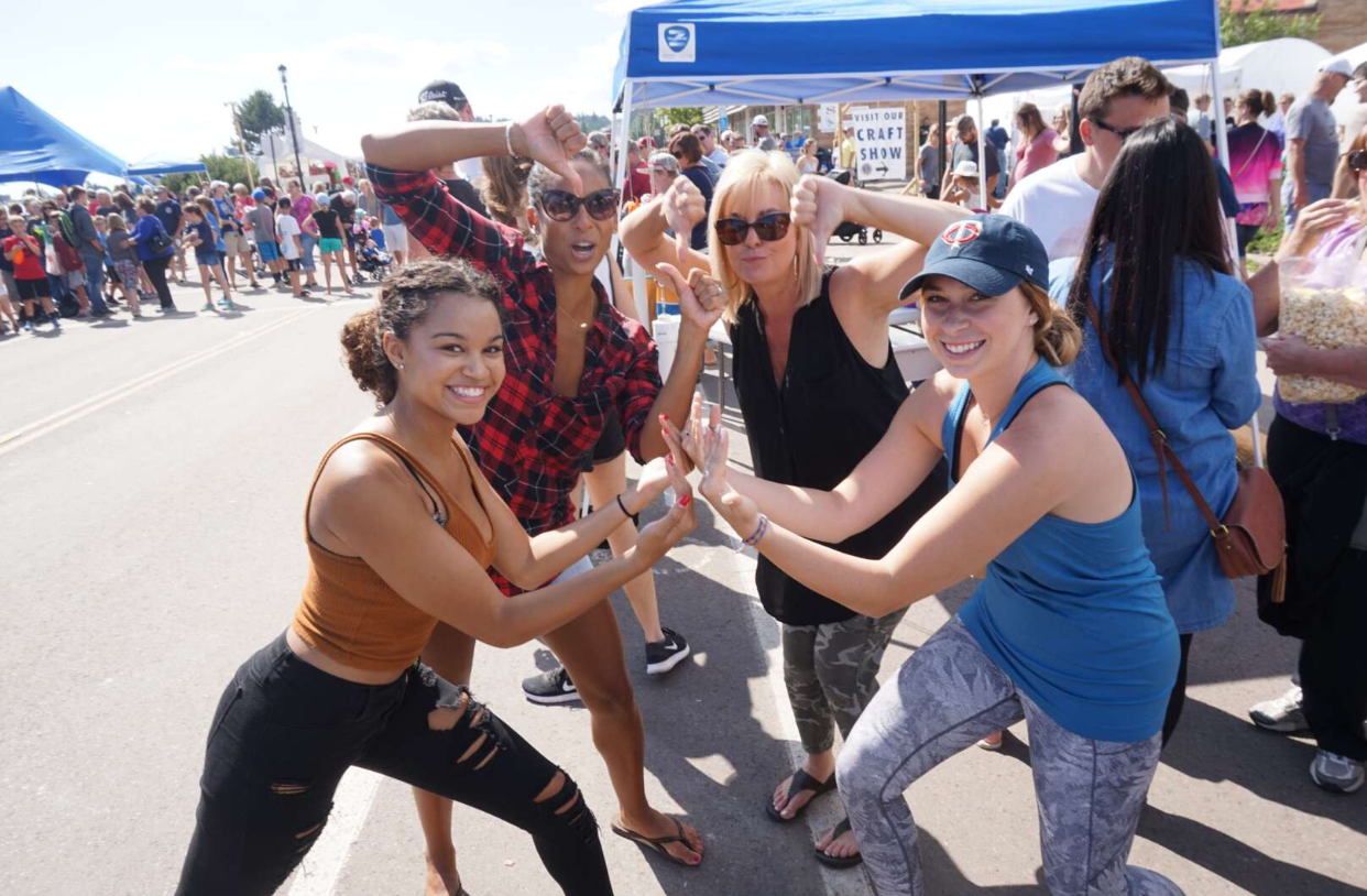 Four women posing playfully and smiling at an outdoor event booth with a crowd in the background.