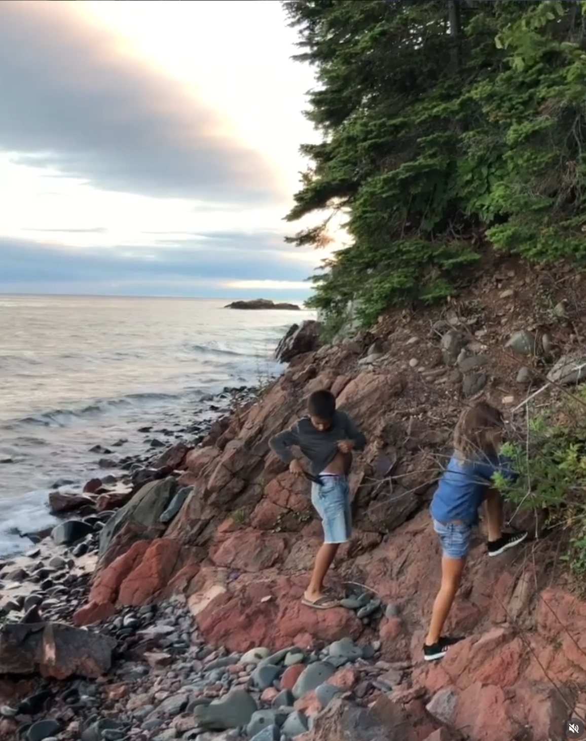 Two boys climbing on a rocky shoreline with trees and the ocean in the background during sunset.