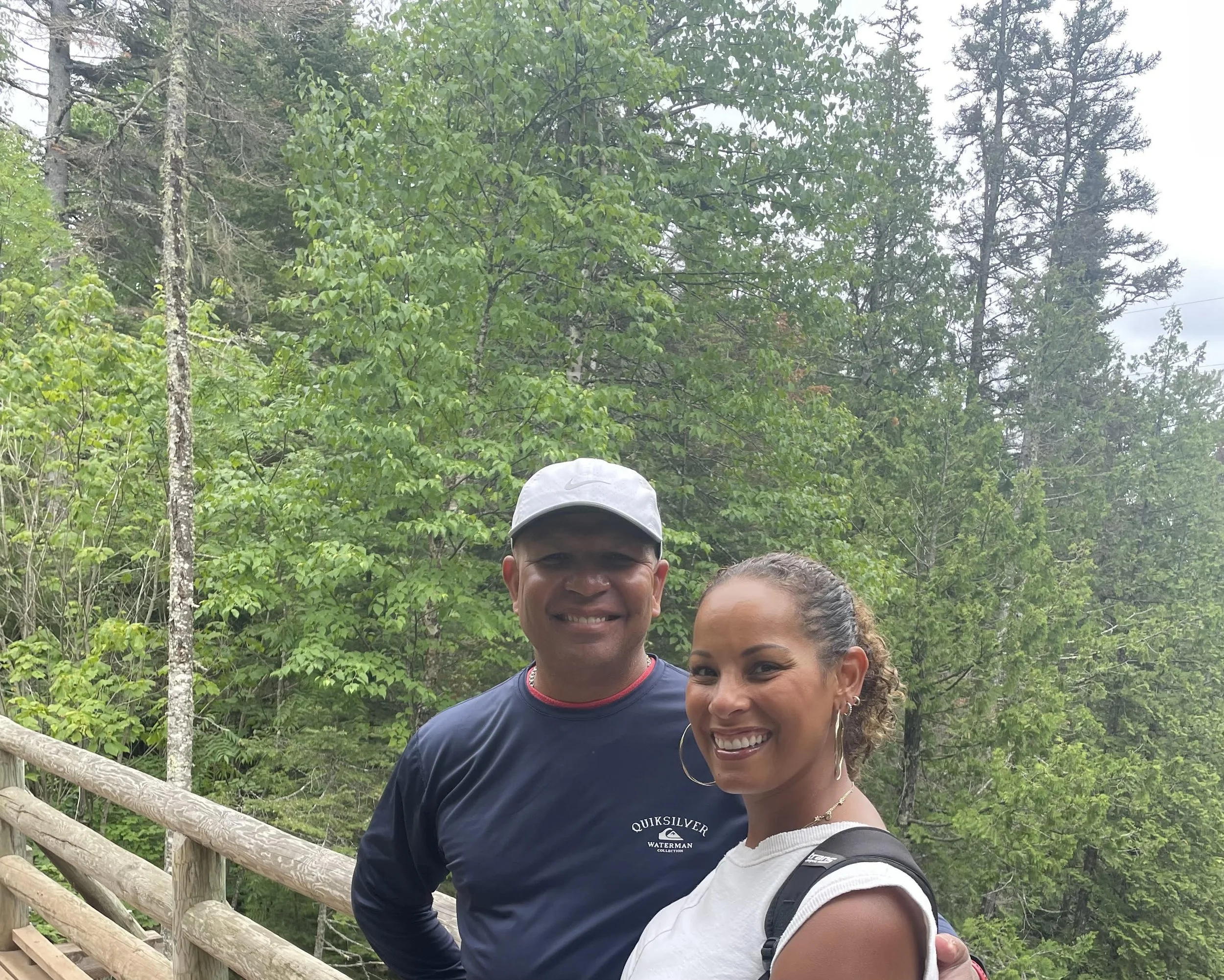 A man and a woman smiling on a wooded trail with green trees in the background.