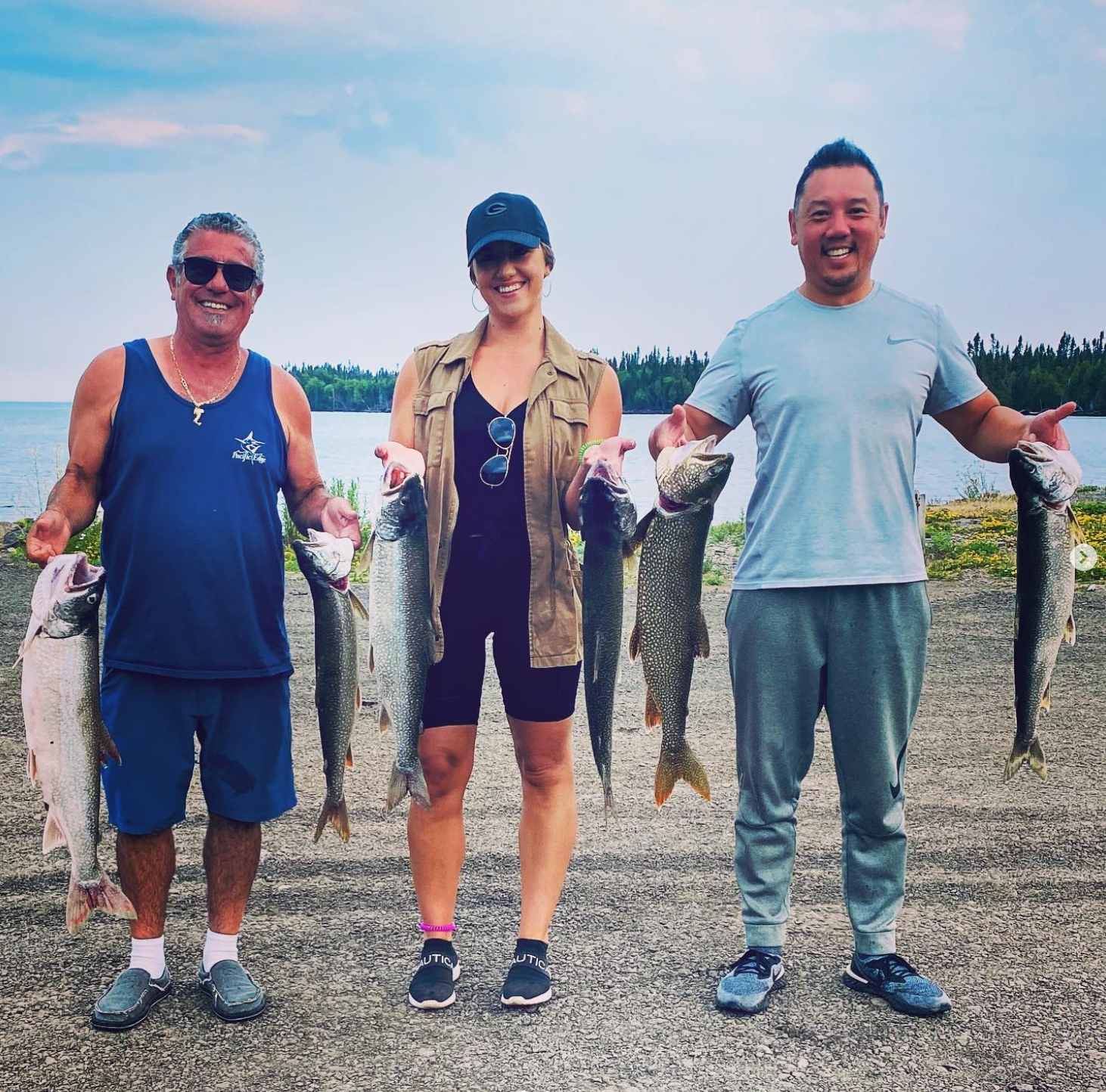 Three people standing outdoors by a lake, holding large fish they caught while fishing.