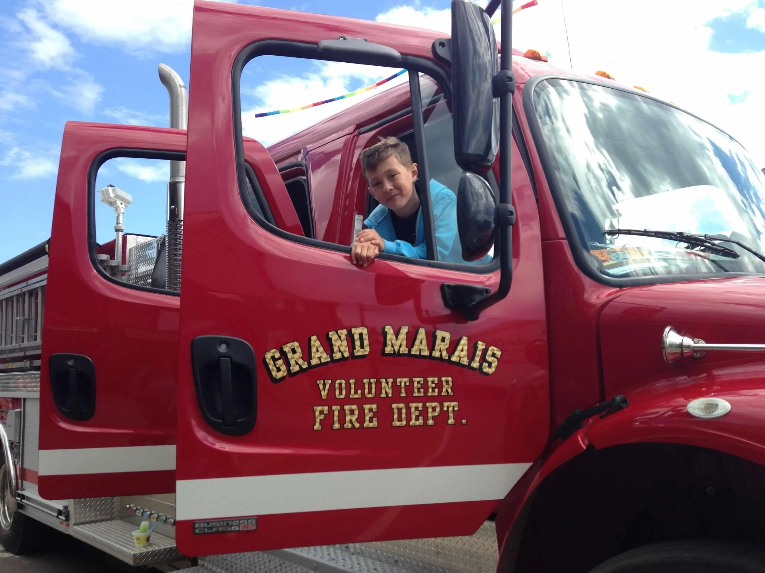 A boy leaning out of the window of a red fire truck labeled Grand Marias Volunteer Fire Dept., with a blue sky and clouds in the background.