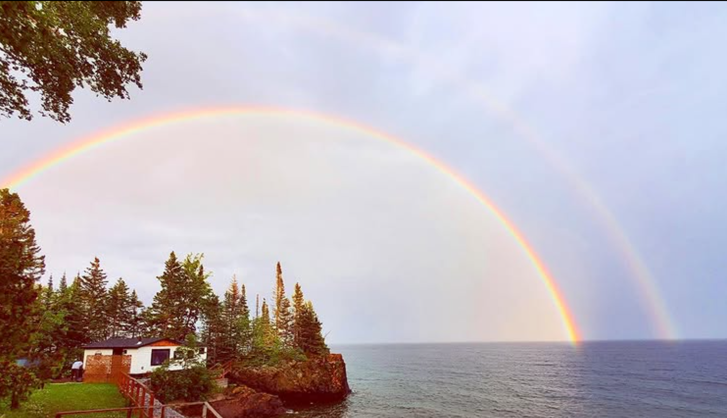 A double rainbow over a body of water near a rocky shoreline with trees and a small building.