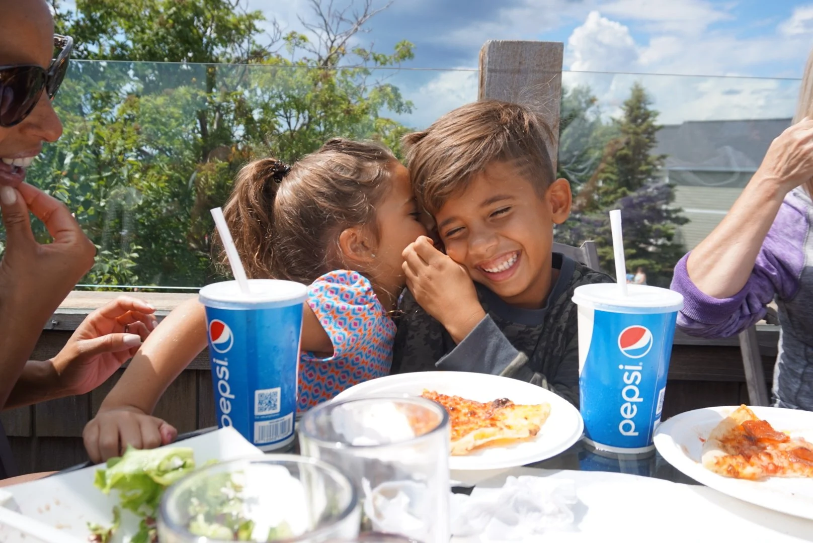 A group of people enjoying a meal outdoors, with two children sharing a laugh. There are pizza slices and Pepsi drinks on the table. The background shows trees and a cloudy sky.