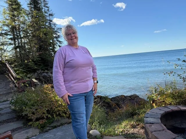 Older woman with white hair smiling, wearing a pastel purple long-sleeve shirt and jeans, standing outdoors near a lake with rocks, trees, and a partly cloudy sky in the background.
