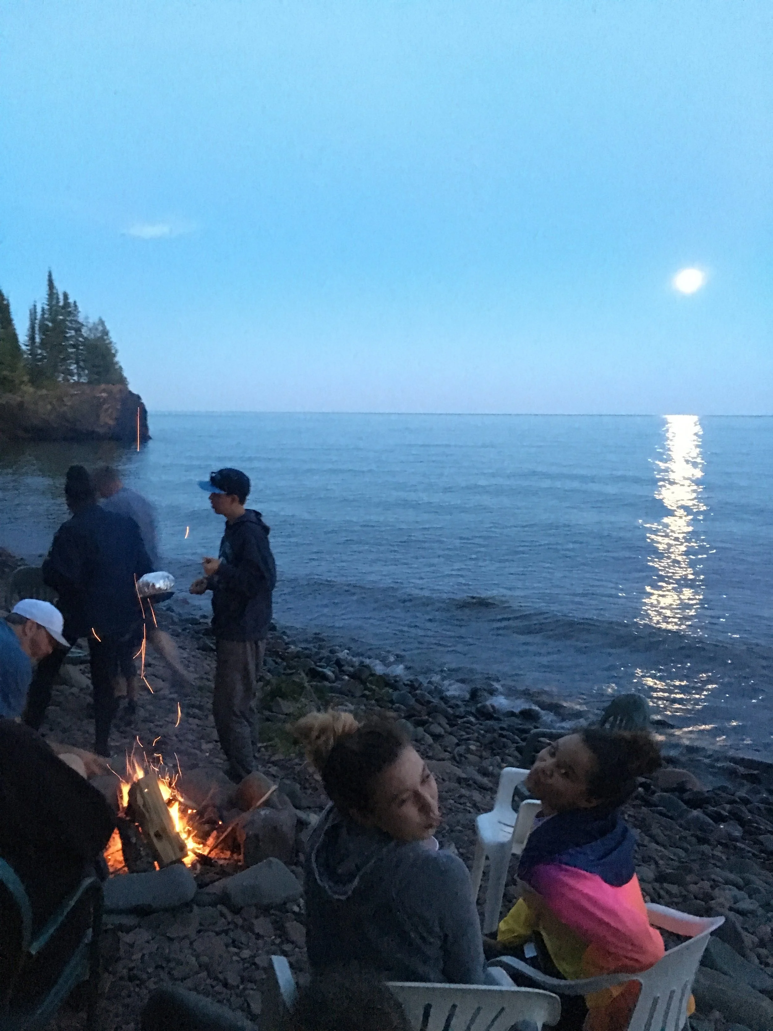 People gathered on a rocky beach by the water at dusk, some sitting around a campfire, with a full moon reflecting on the water, in a scenic outdoor setting.