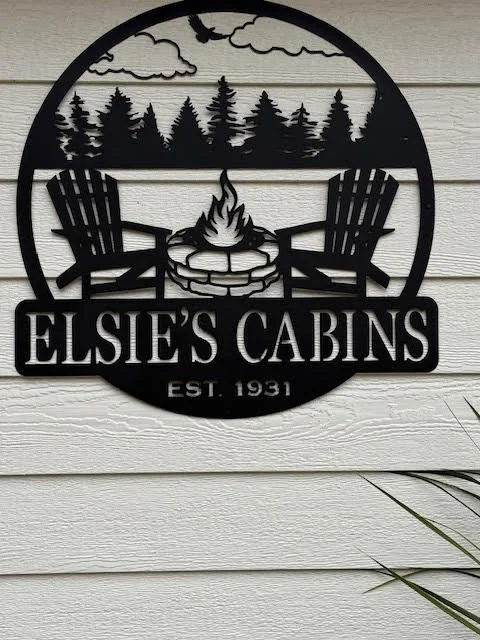 Metal sign with a circular design featuring two Adirondack chairs, a campfire, pine trees, clouds, and the text 'Elsie's Cabins, Est. 1931', mounted on a white wooden wall.