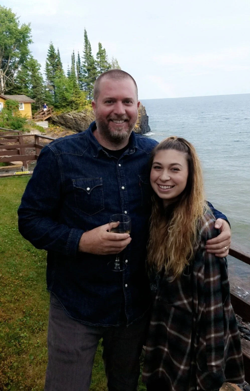 A man with a beard and a woman with long wavy hair standing outdoors near water, smiling with the ocean and trees in the background, holding a glass of wine.
