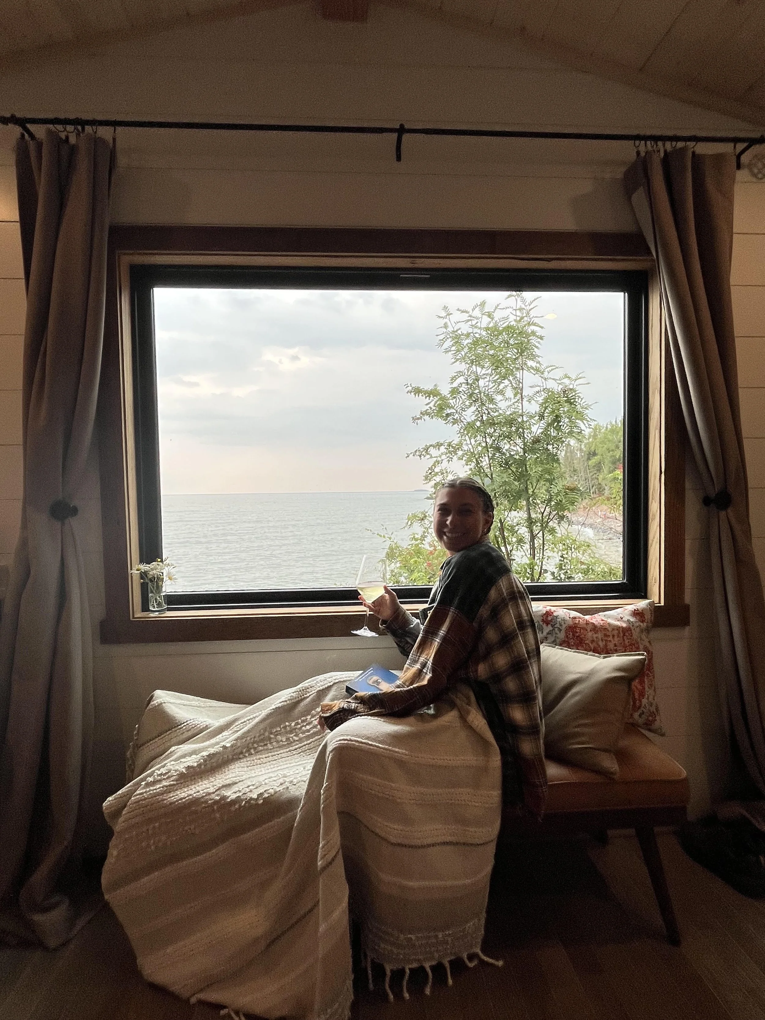 A woman sitting on a bed in front of a window overlooking a lake, holding a glass of drink, wearing a plaid shirt and smiling.