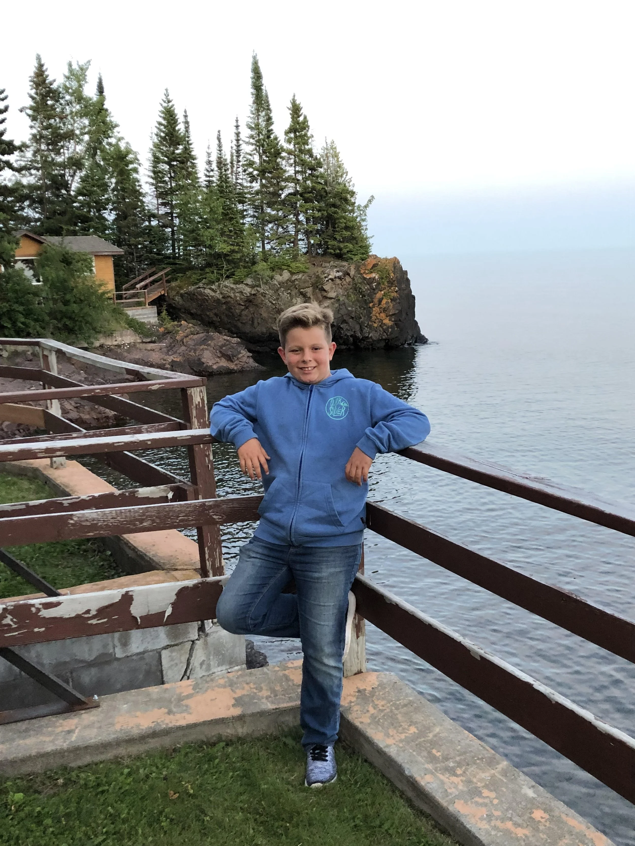 A young boy in a blue hoodie and jeans standing by a wooden railing along the water's edge, with trees and a rocky shoreline in the background.