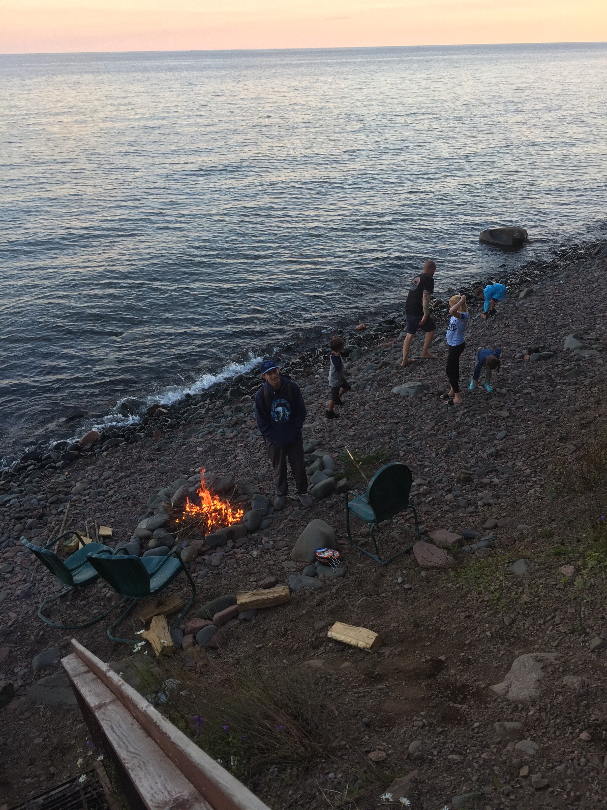 People, including children and an adult, on a rocky beach at sunset, with some gathered around a small campfire and others exploring near the water, with chairs and logs nearby.