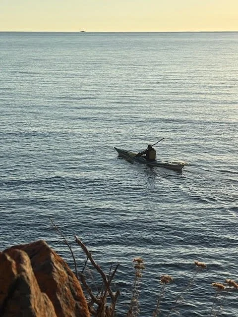 A person kayaking on calm water near a rocky shoreline at sunset.