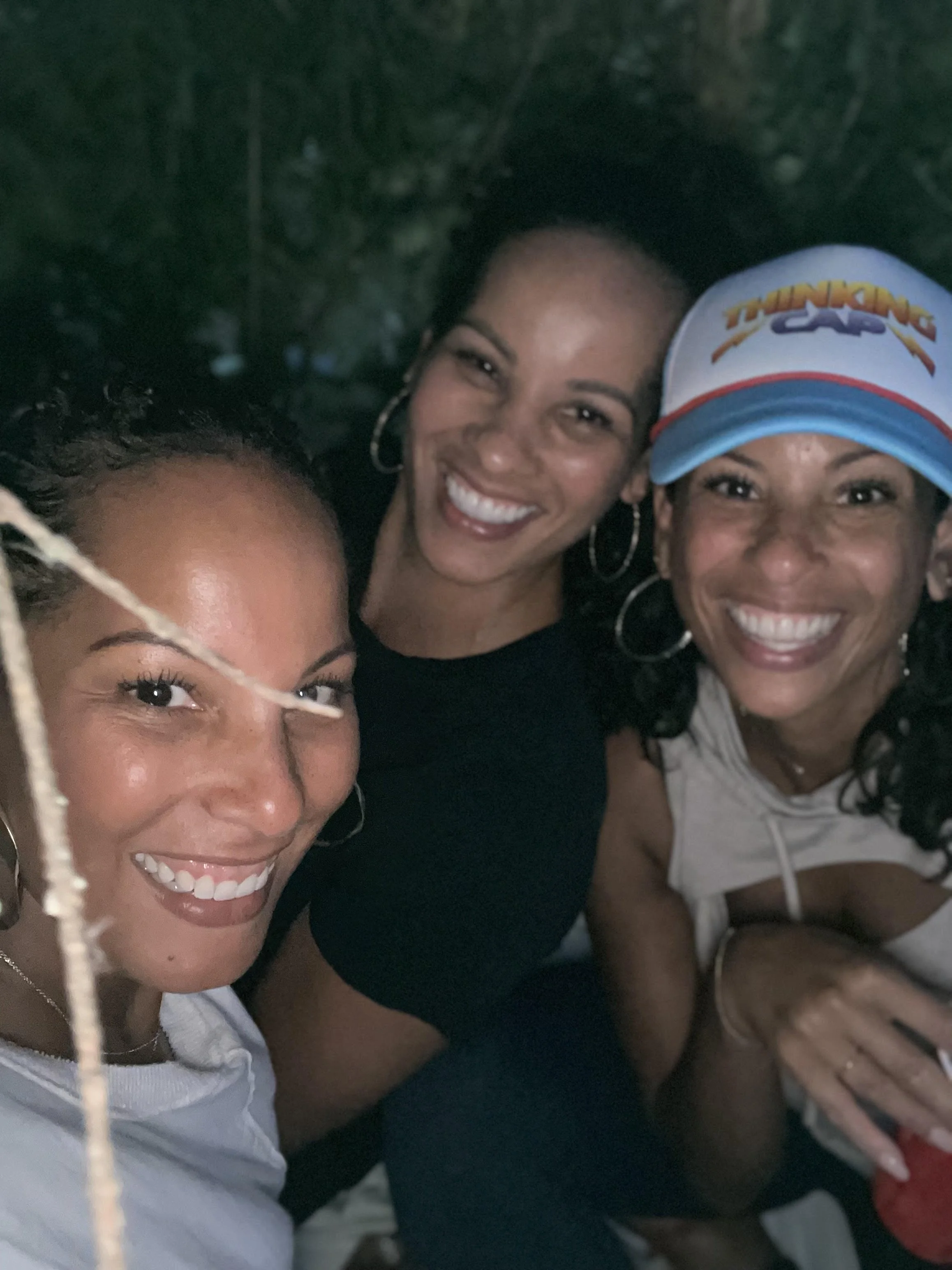 Three women smiling and posing for a selfie outdoors, one wearing a Thundering Cack cap.