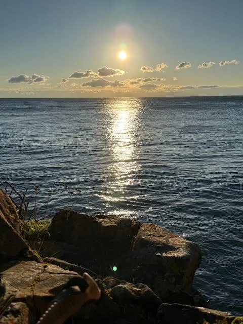 Sun setting over the ocean with reflected sunlight on the water, rocky shoreline in the foreground, partly cloudy sky.