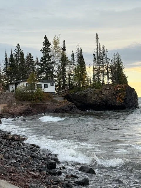 A small house on a rocky shoreline with tall pine trees and a large rock formation, with water gently lapping against the shore and a cloudy sky overhead.