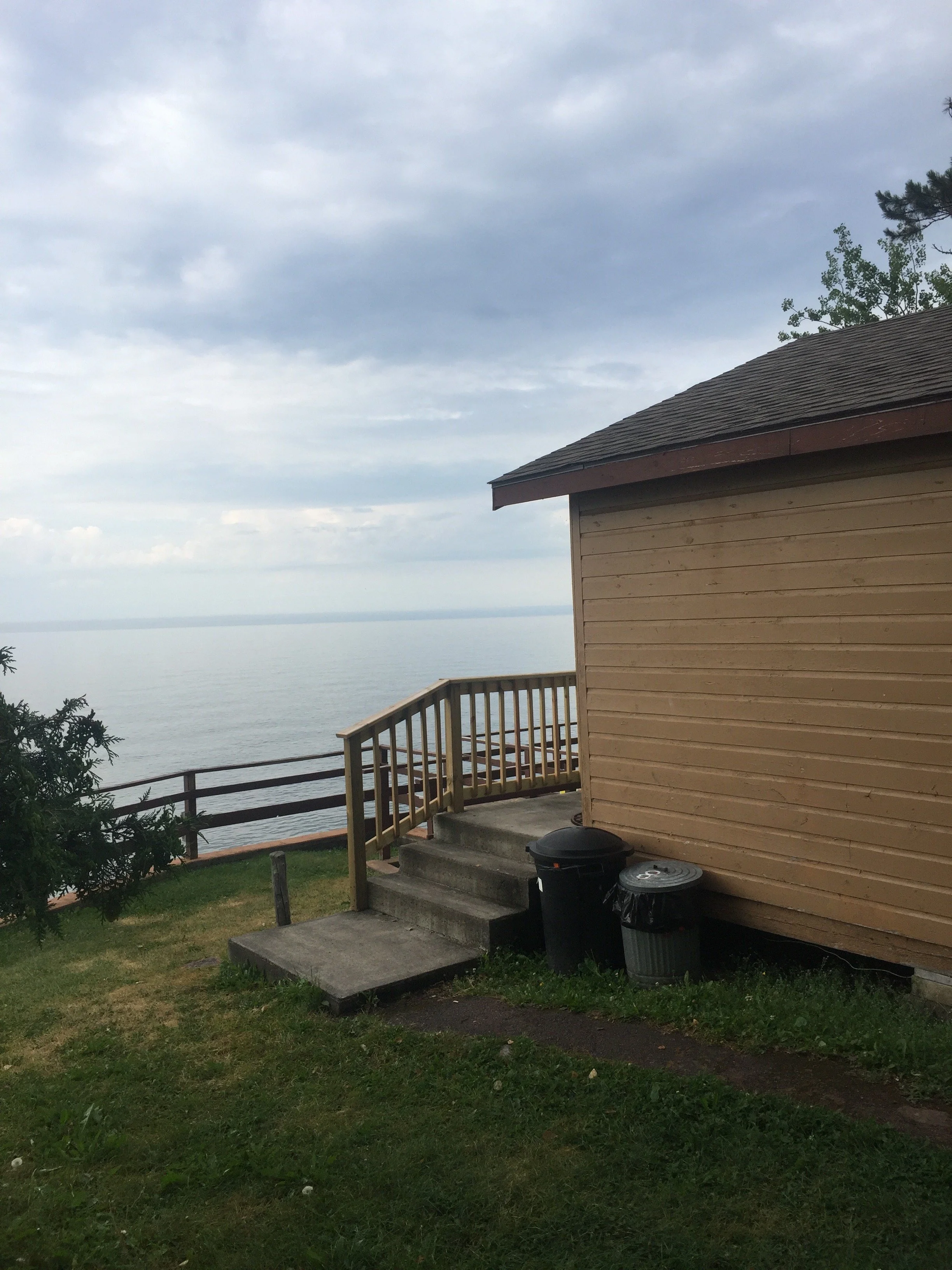 A small house or shed by the water with a wooden deck and stairs leading down to the grassy yard, three trash cans near the house, and a cloudy sky over a calm body of water in the background.