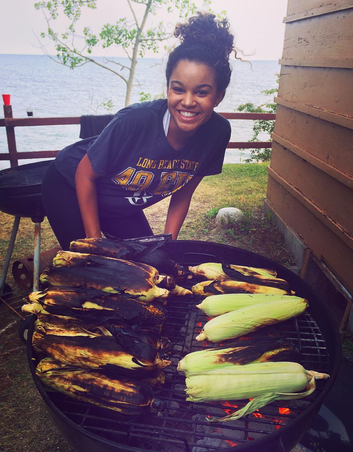 A young woman grilling corn on a barbecue with a lake and trees in the background.