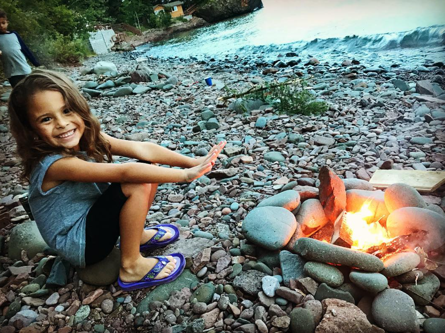 A young girl sitting on a rock on a rocky beach, smiling and reaching towards a small campfire surrounded by stones, near a body of water with trees and houses in the background.