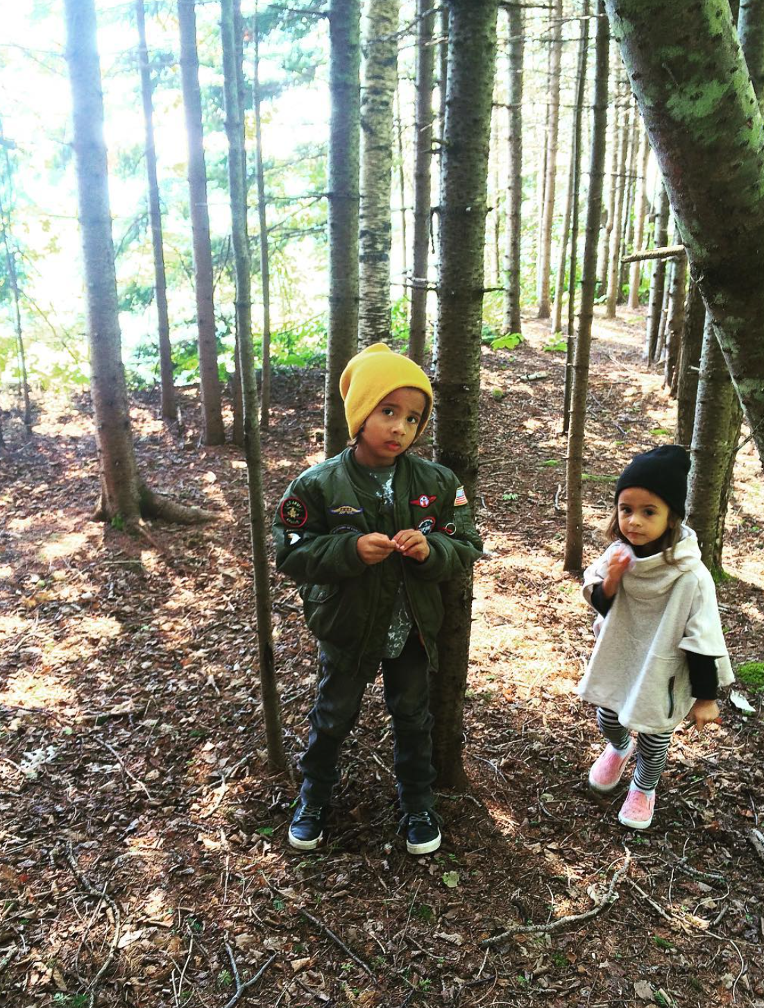 Two kids standing in a wooded forest with tall trees, one wearing a yellow beanie and green jacket, the other wearing a black beanie with a light jacket, and striped leggings.