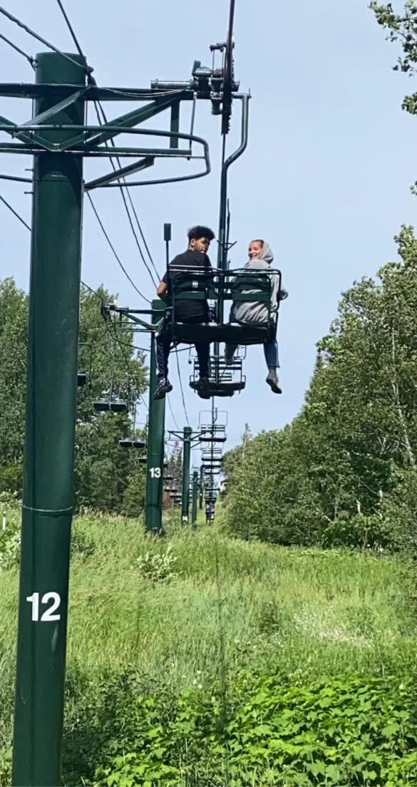 Two people riding a ski lift in a green, forested area on a sunny day, talking to each other.