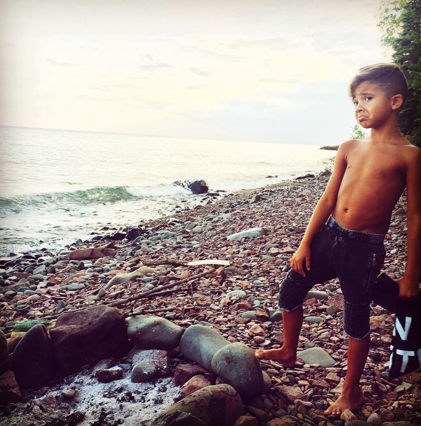 A shirtless boy with dark shorts standing on a rocky beach with water and trees in the background.