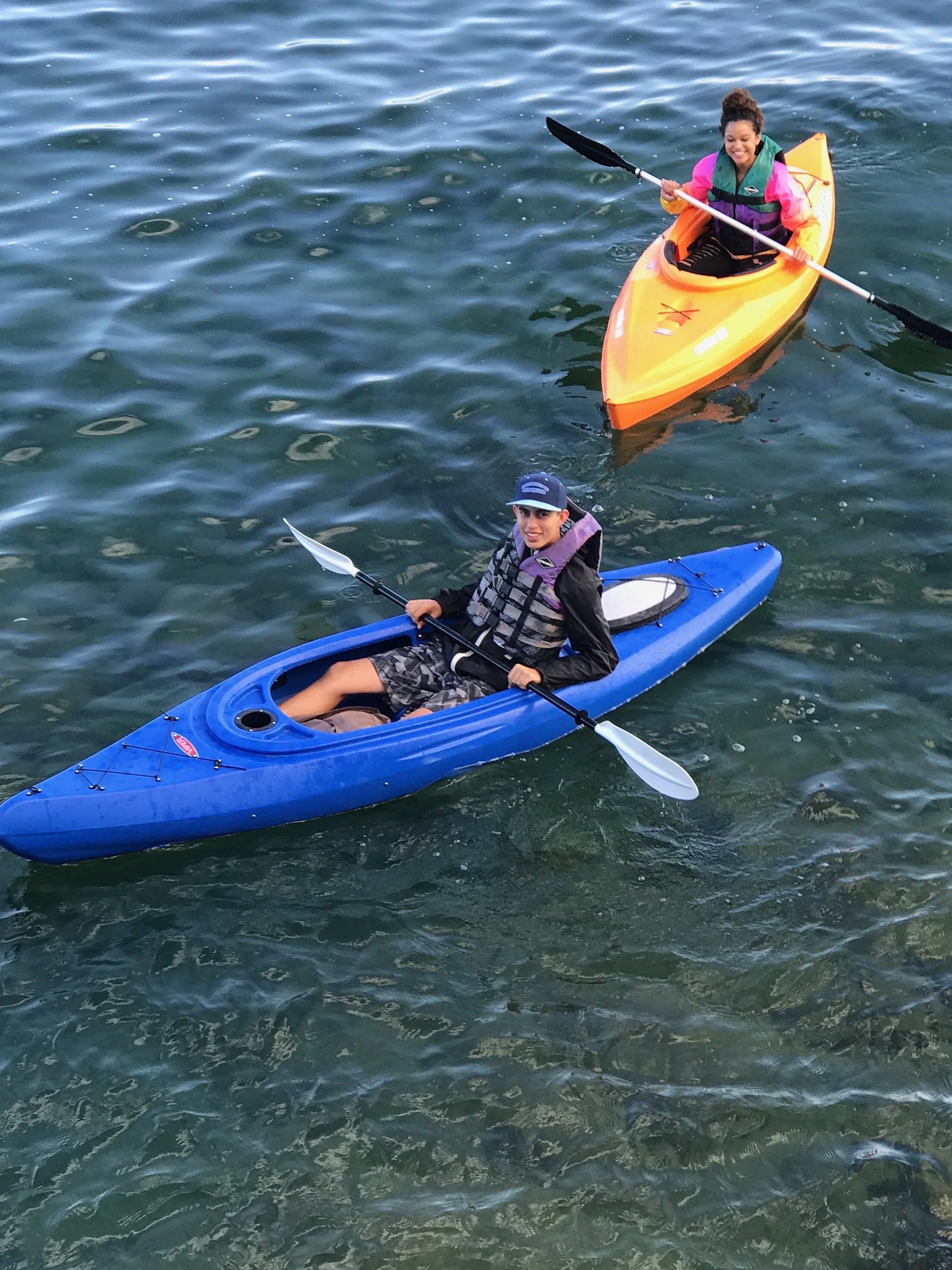 Two people kayaking in a body of water, one woman in an orange kayak and a young man in a blue kayak, both smiling and holding paddles.