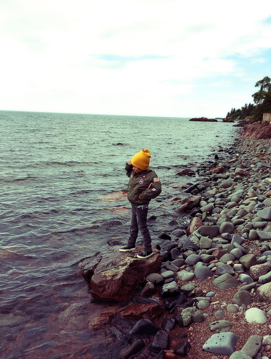 A young boy wearing a yellow beanie, green jacket, black pants, and sneakers standing on a large rock at the edge of a rocky shoreline, looking down at the water with his hand on his head, overlooking a body of water with a distant island and trees.