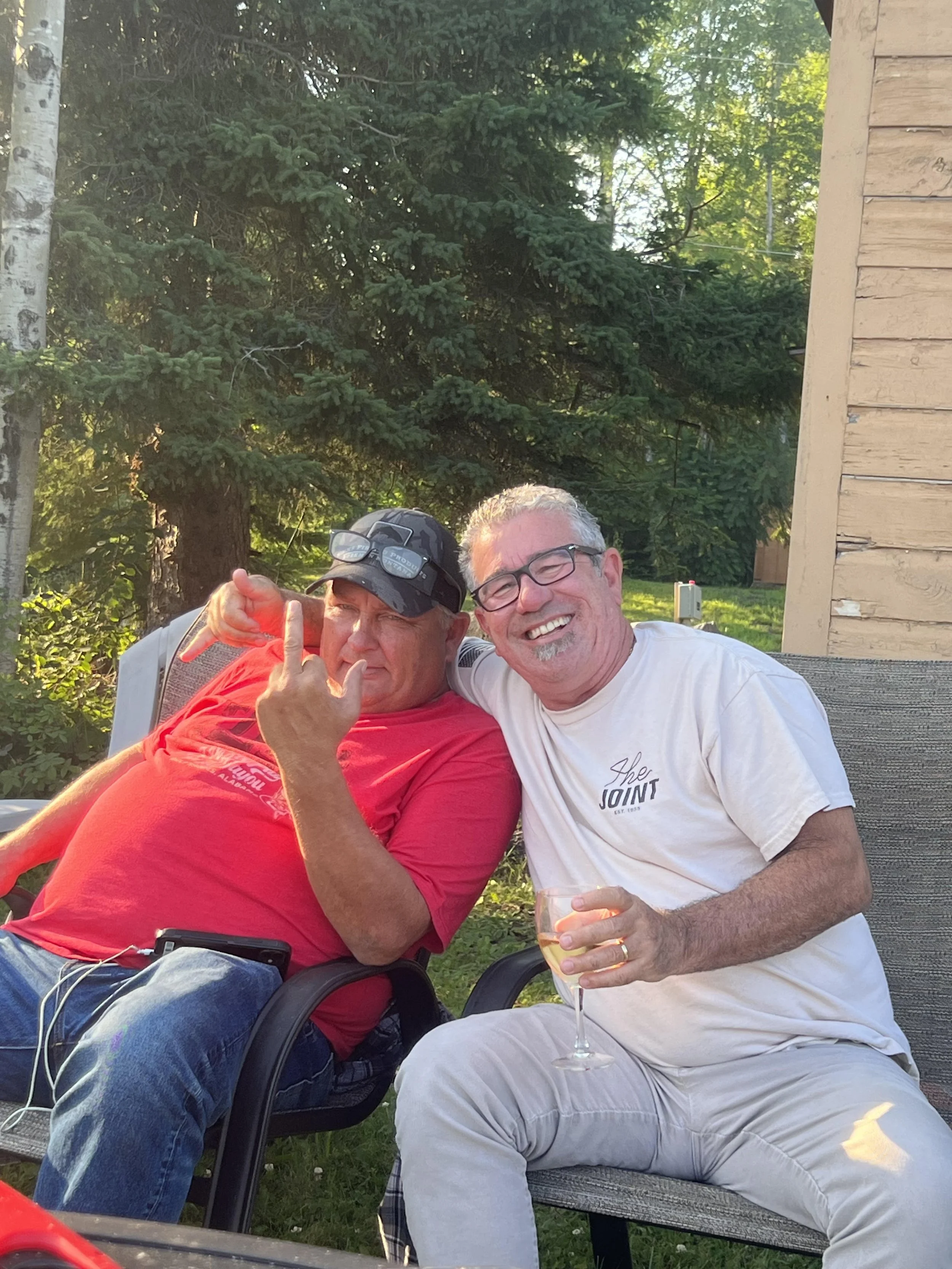 Two middle-aged men sitting outdoors on a bench, smiling and enjoying drinks, with a background of green trees and a wooden building.