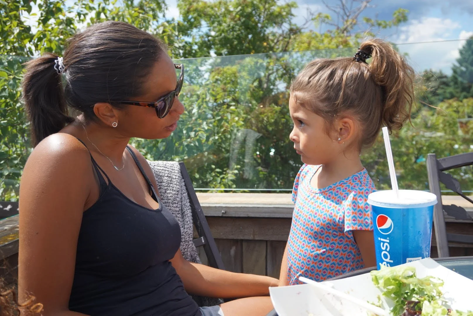 A woman and a young girl sit facing each other outdoors, engaged in conversation. The woman wears sunglasses and a black tank top, while the girl wears a colorful patterned shirt. There is a large Pepsi cup and food on the table in front of them, wit