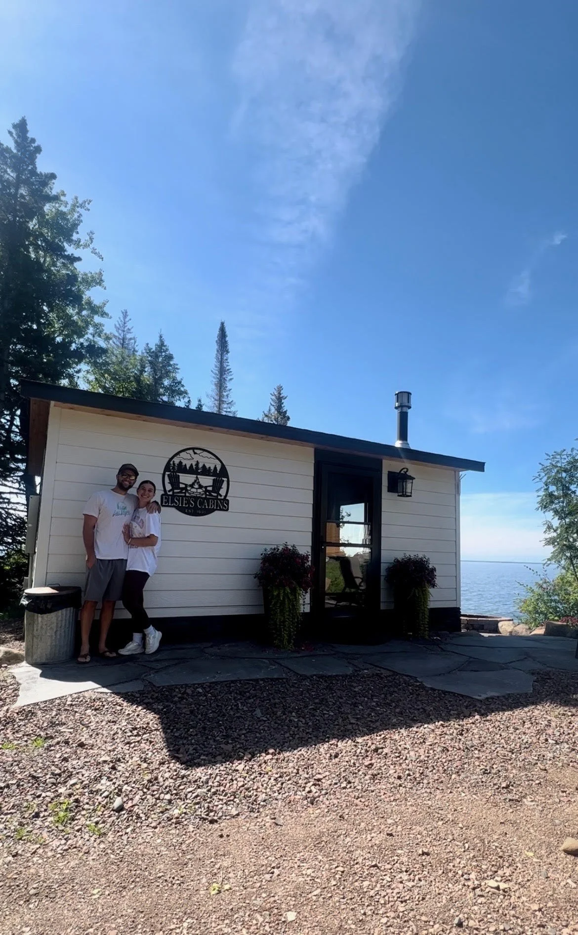 A small cabin by the water with a sign reading Elsie's Cabins, two people standing in front smiling, trees around, and a view of the water in the background on a sunny day.
