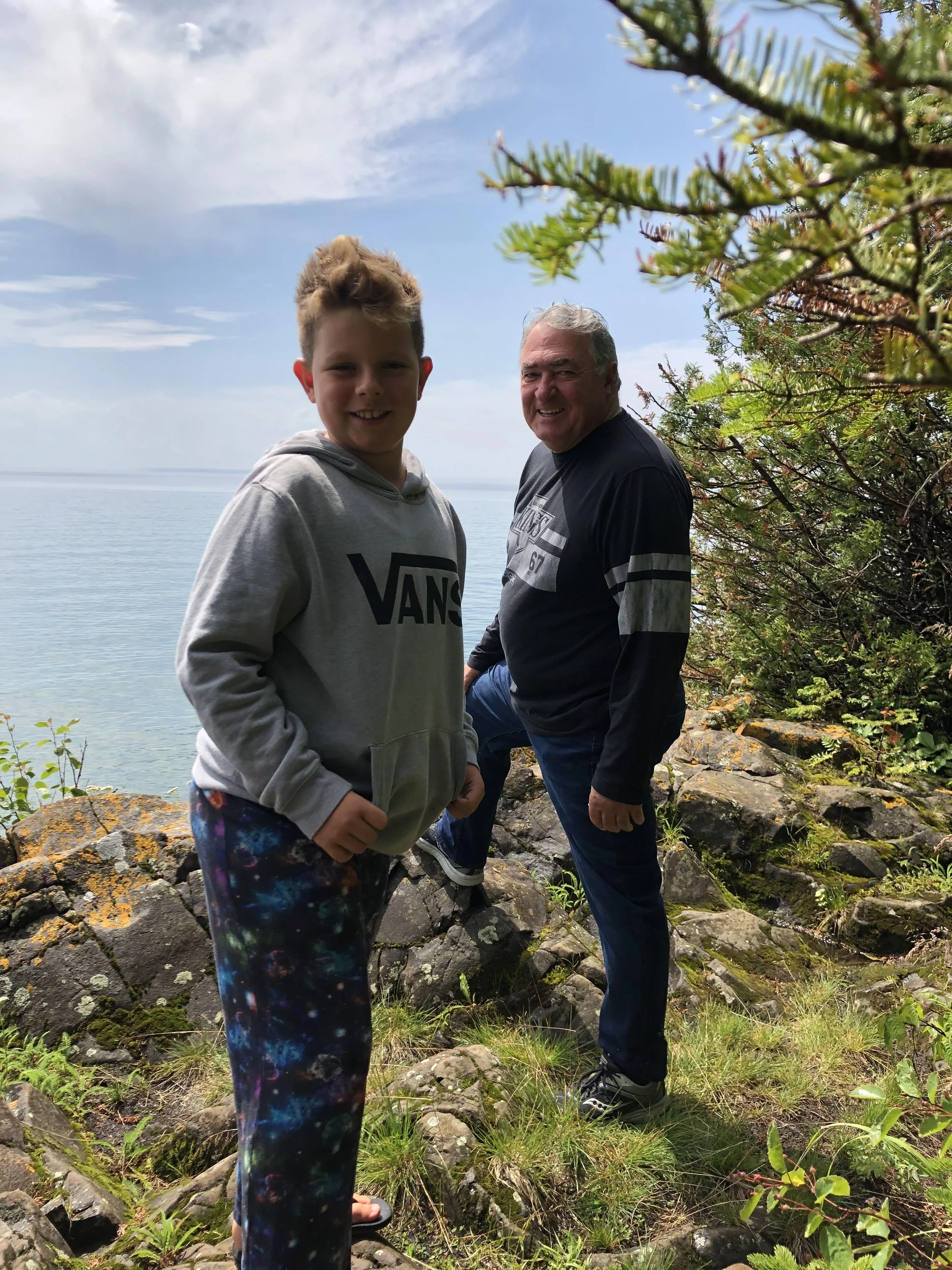 A young boy and an older man standing outdoors on rocky terrain near a body of water, with trees and a partly cloudy sky in the background. The boy is smiling and wearing a grey hoodie with 'VANS' on it and colorful galaxy-patterned pants. The man is