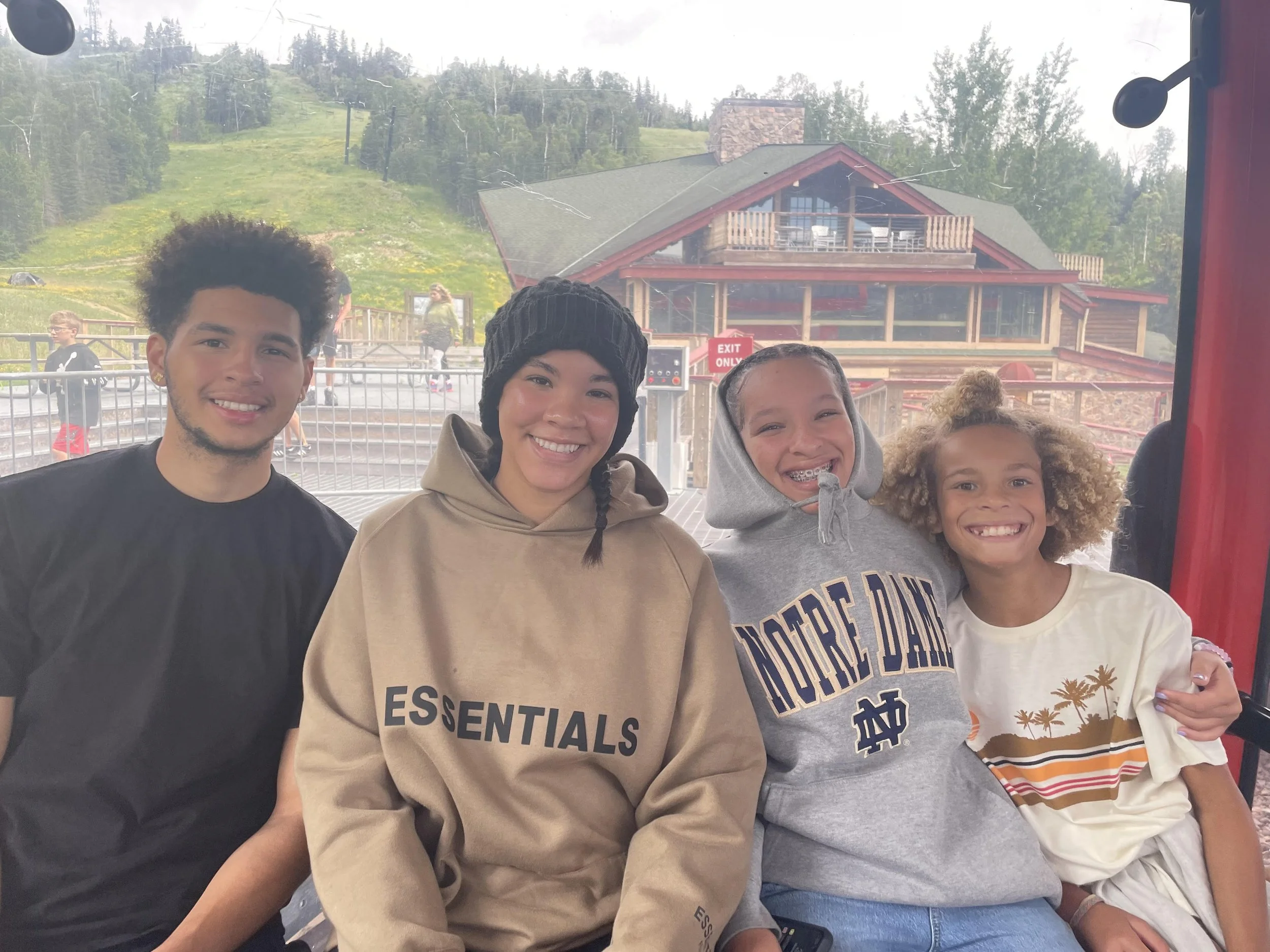 Four children smiling on a Ferris wheel with a mountainous landscape and a building in the background.