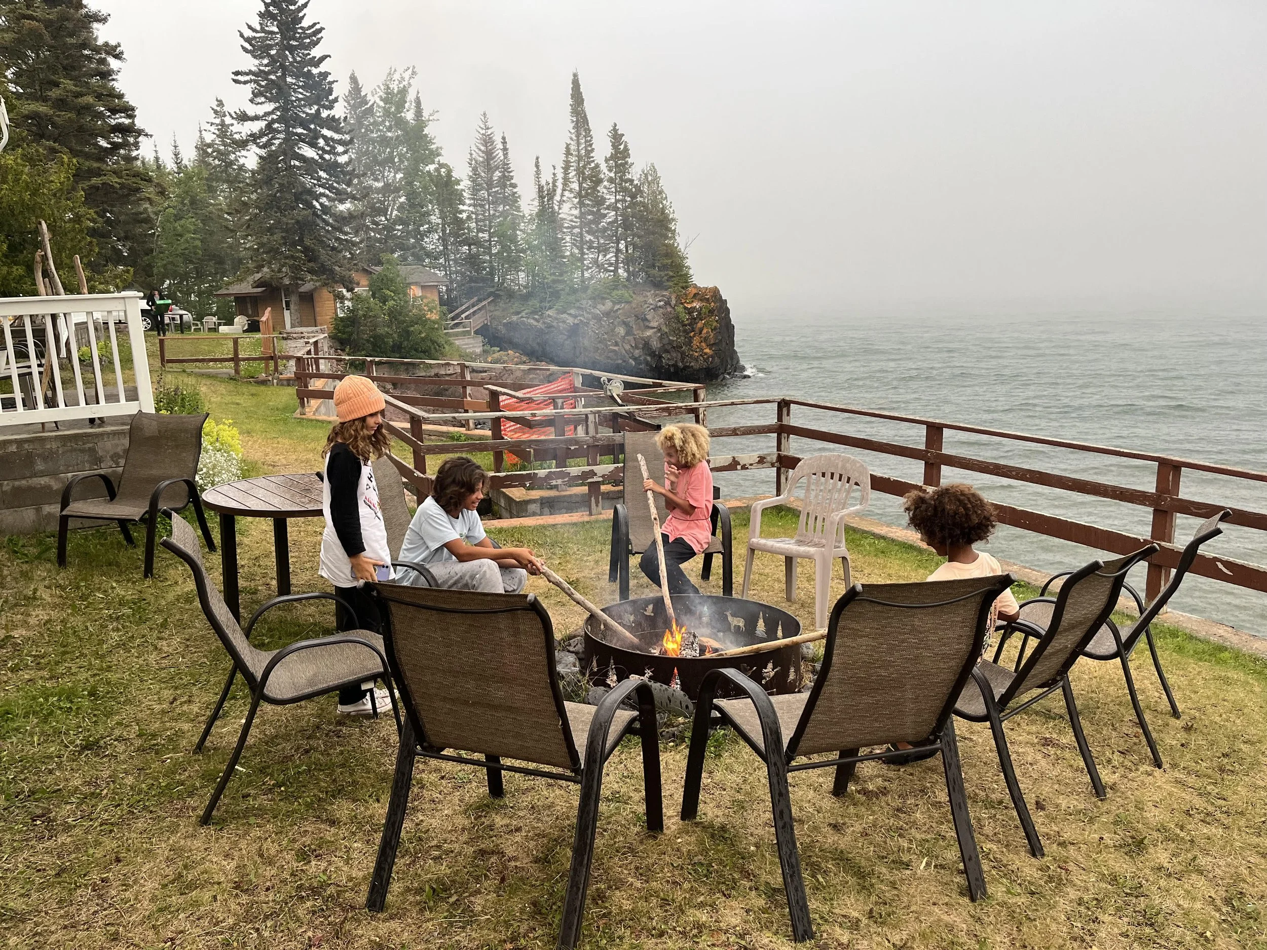 Children sitting and standing around a campfire on a grassy area near the water with a wooden railing, trees, and small houses in the background.