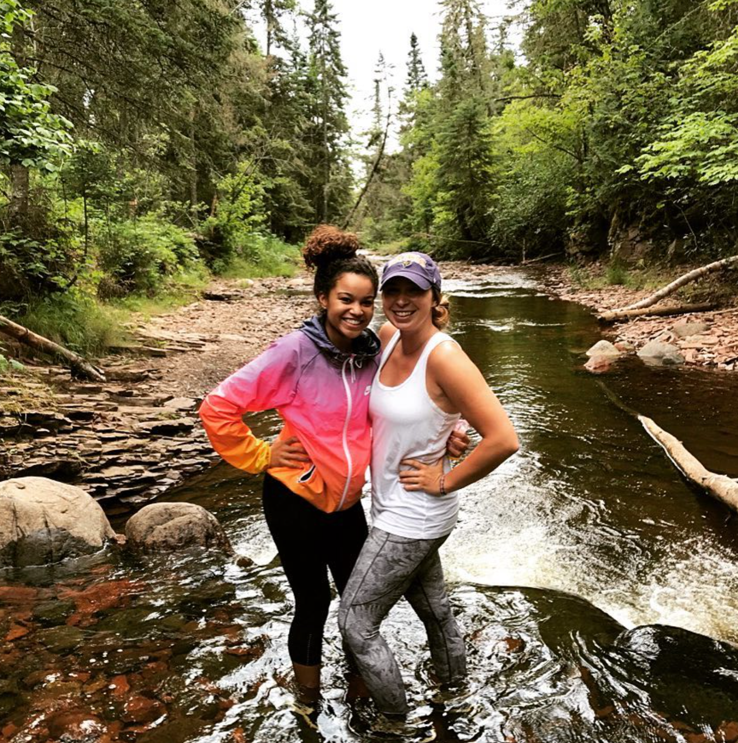 Two women smiling and posing in a shallow river surrounded by trees and rocks in a forest