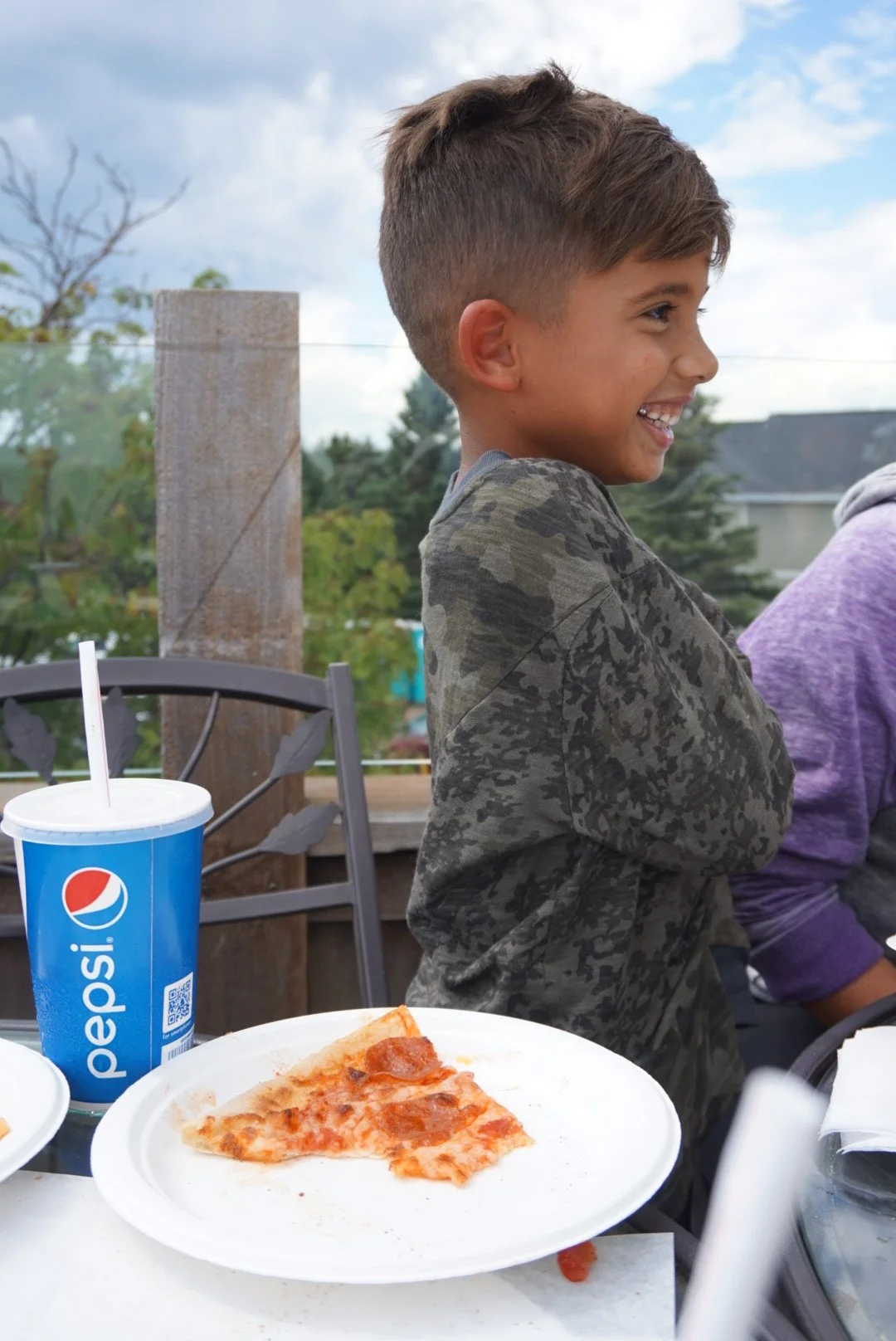 A young boy laughing outdoors at a table with a slice of pepperoni pizza on a white paper plate and a Pepsi drink in a blue cup, with a partly cloudy sky in the background.