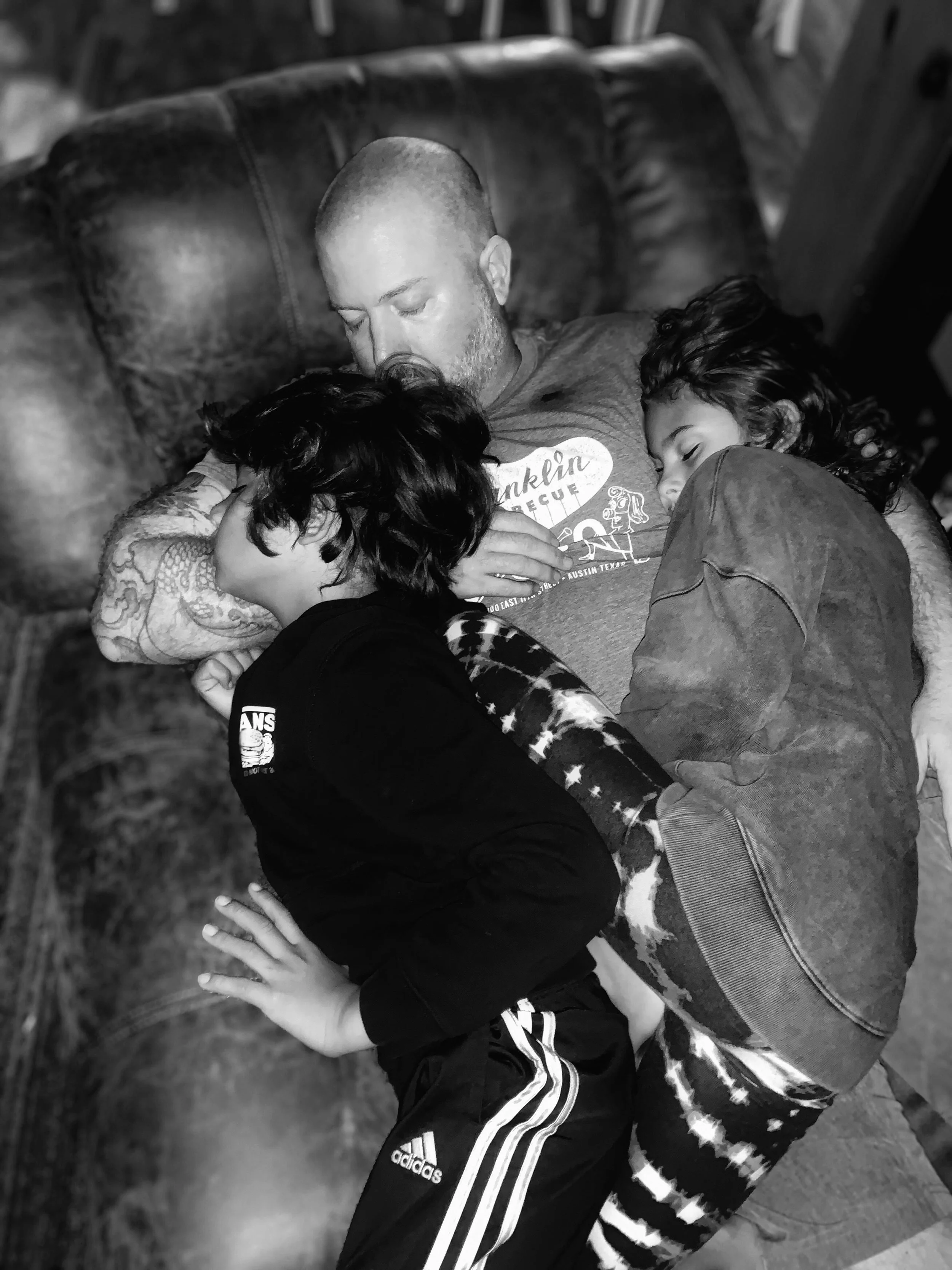 A man lying on a leather couch, asleep, with two children cuddled on either side of him.