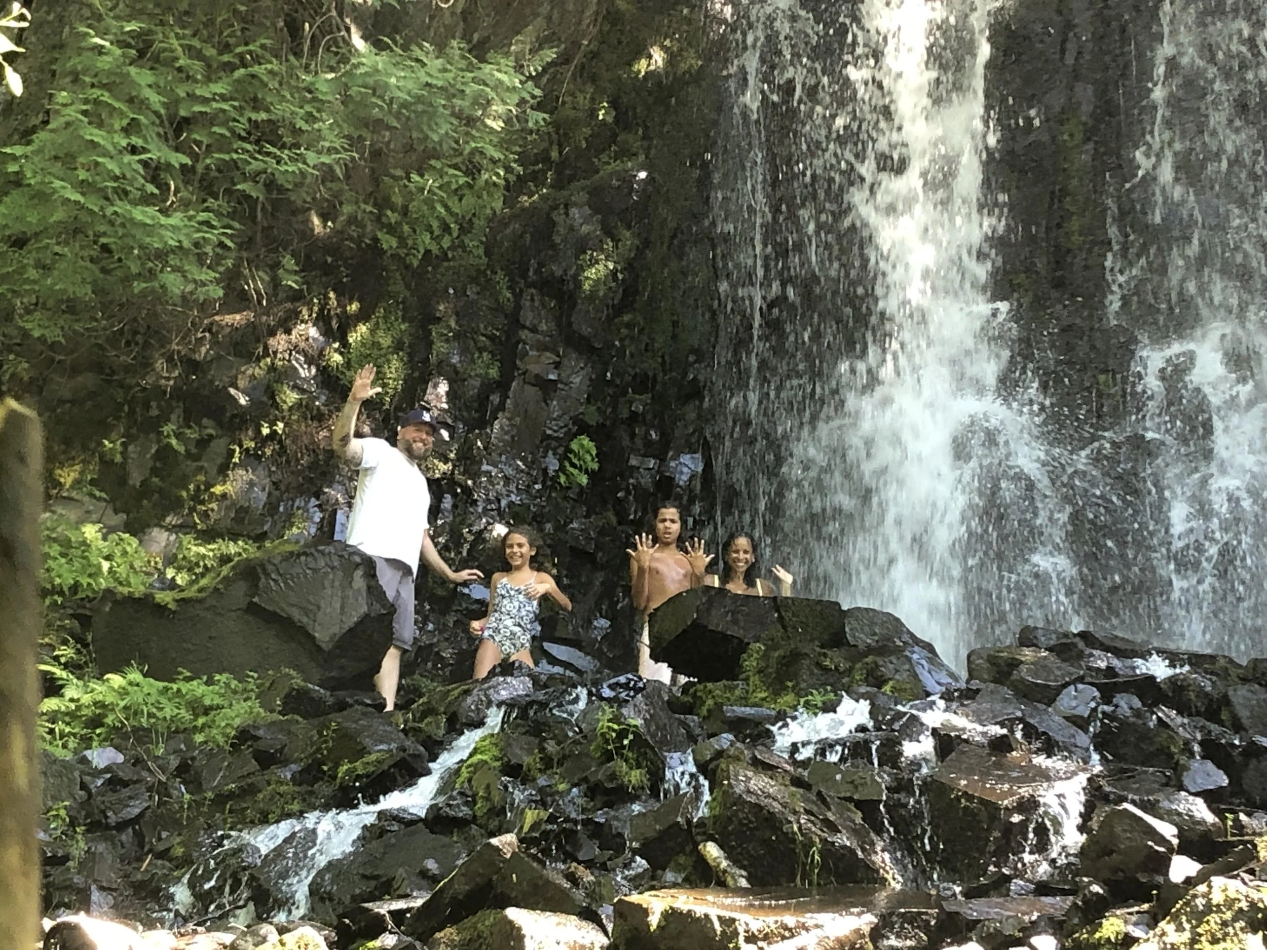 Four people standing on rocks in front of a waterfall in a forest, smiling and waving, during daytime.