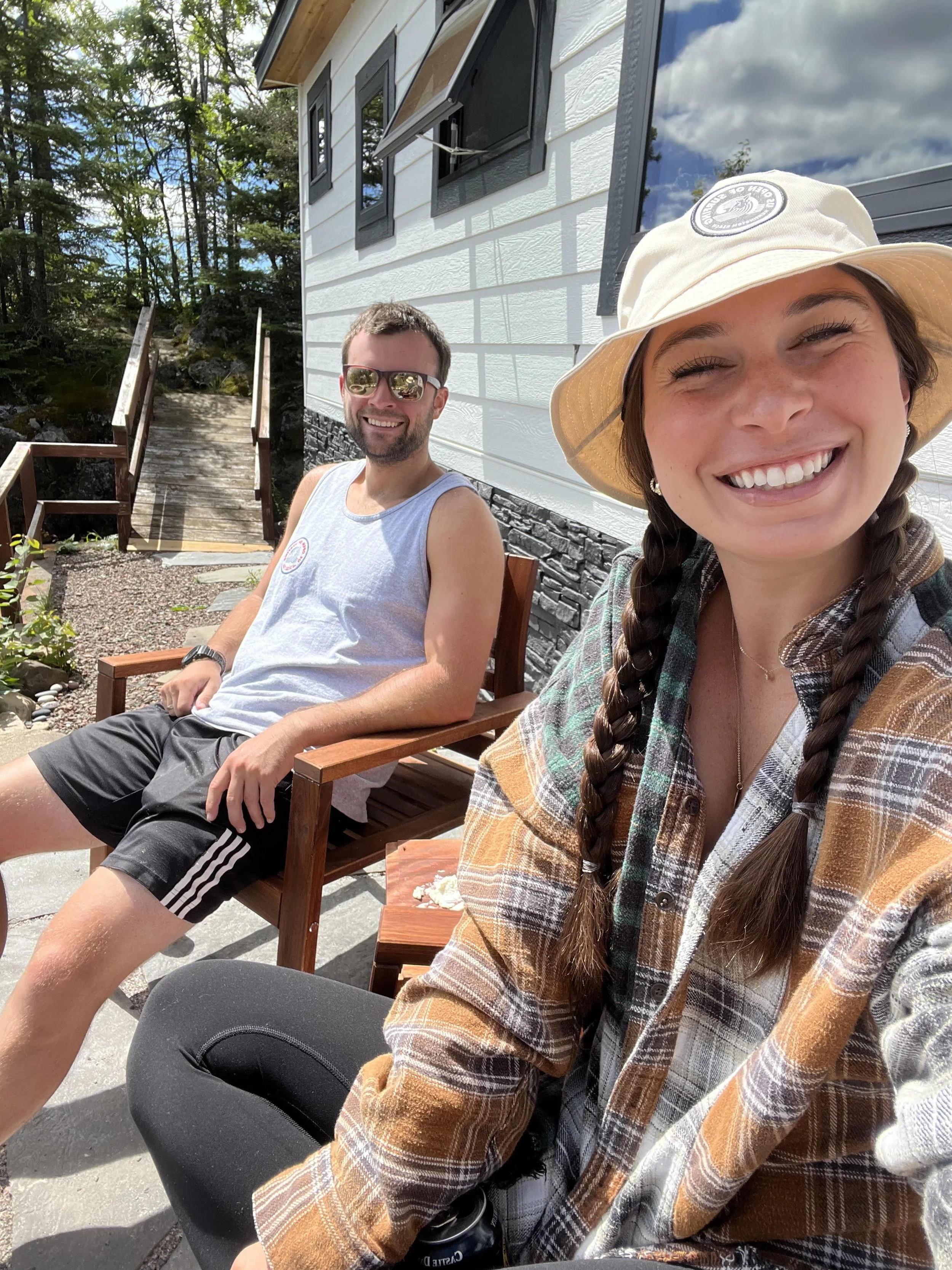 A young woman with braided hair, wearing a wide-brimmed hat and plaid shirt, smiling for a selfie outdoors, with a man sitting on a wooden chair behind her, in front of a house with white siding and black window frames, surrounded by trees and blue s